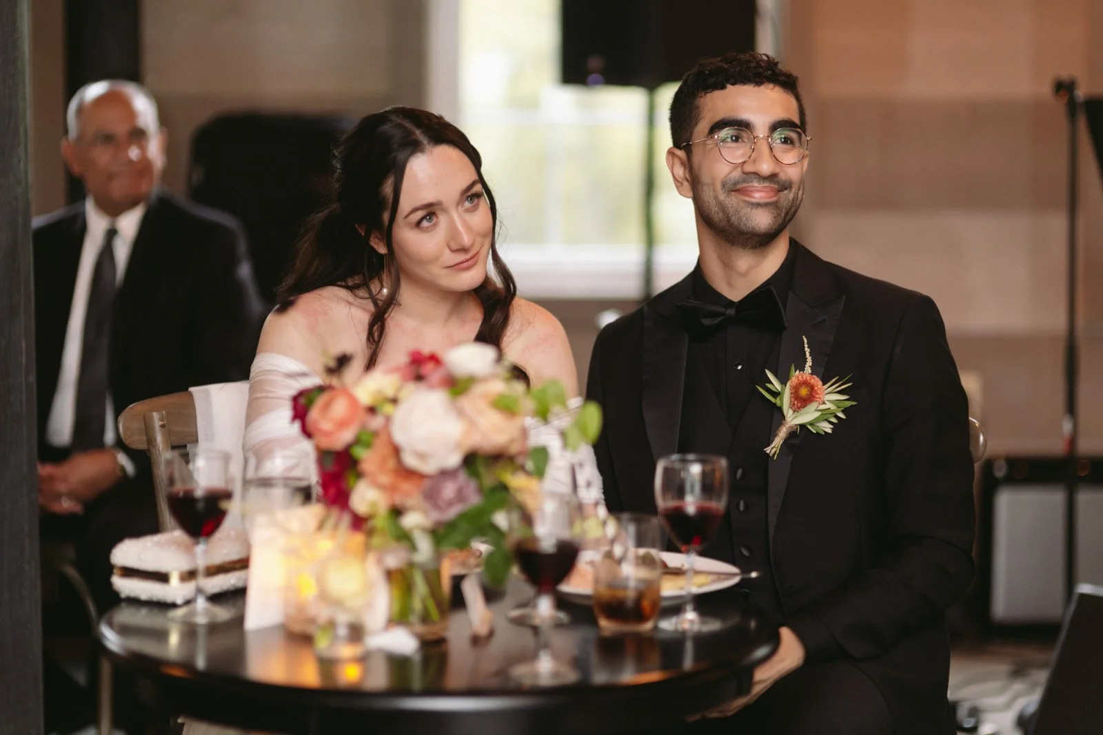 A man and woman in formal attire sitting at a wedding reception table, smiling; the woman has dark hair and a white off-shoulder dress, and the man has glasses and a black tuxedo with a boutonnière; there are wine glasses, a floral centerpiece, and a