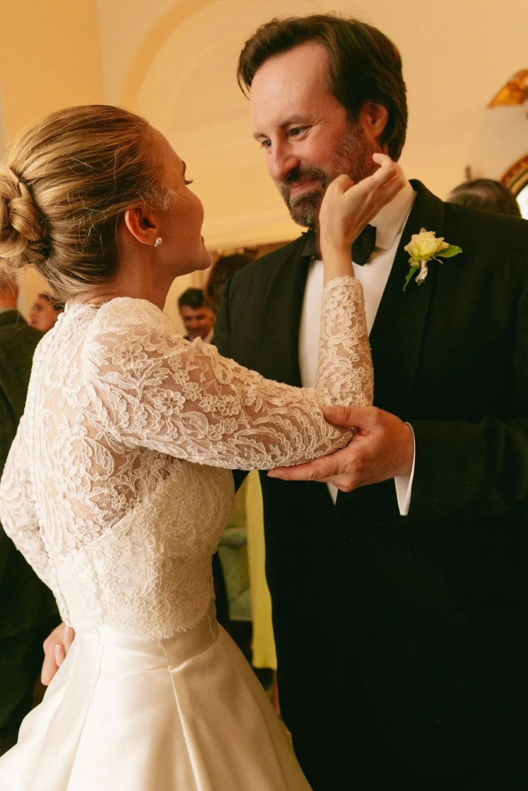 A bride and groom dancing at their wedding reception. The bride is wearing a lace wedding dress and the groom is in a black tuxedo with a white boutonniere. They are looking into each other's eyes, smiling.