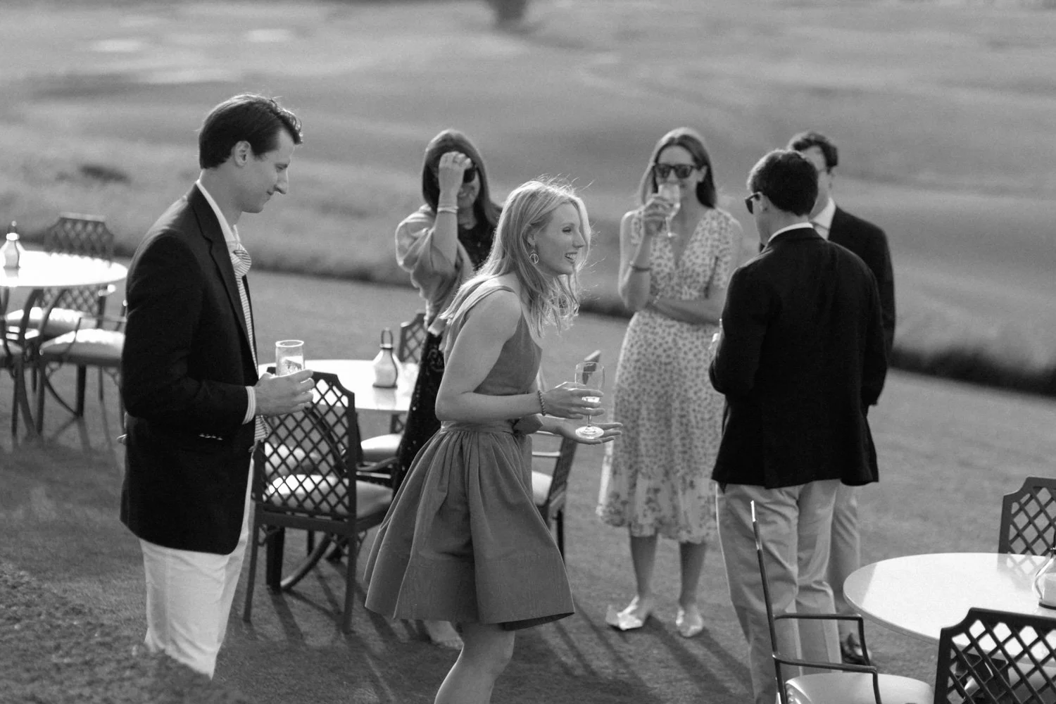 A group of well-dressed people socializing outdoors at a daytime gathering, with some holding drinks and standing near tables.