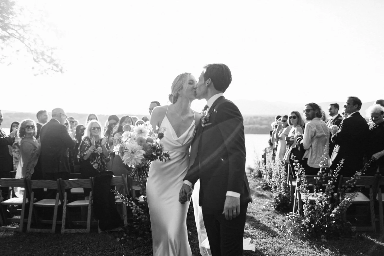Black and white photo of a wedding ceremony outdoors by a river, with a bride and groom kissing, surrounded by guests.