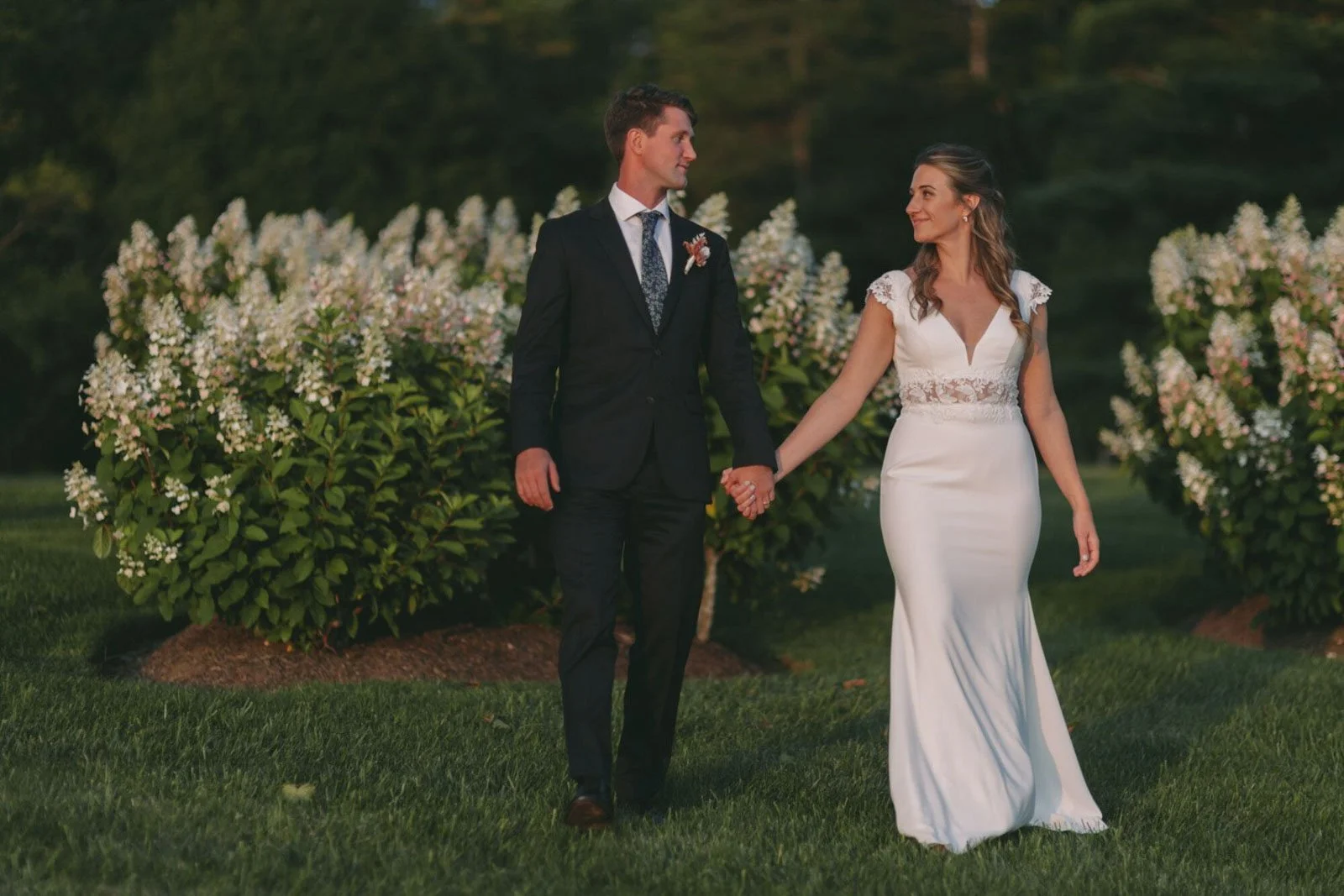 A couple in wedding attire holding hands in a garden with flowering bushes in the background.