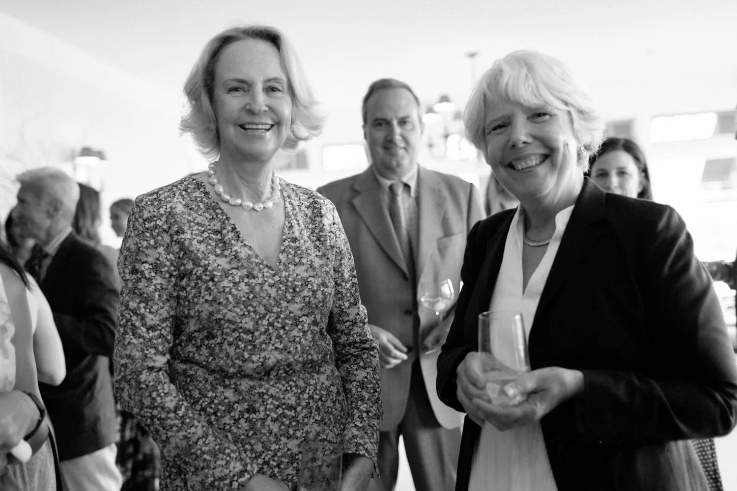 Group of three smiling people at a social gathering, with additional people in the background.