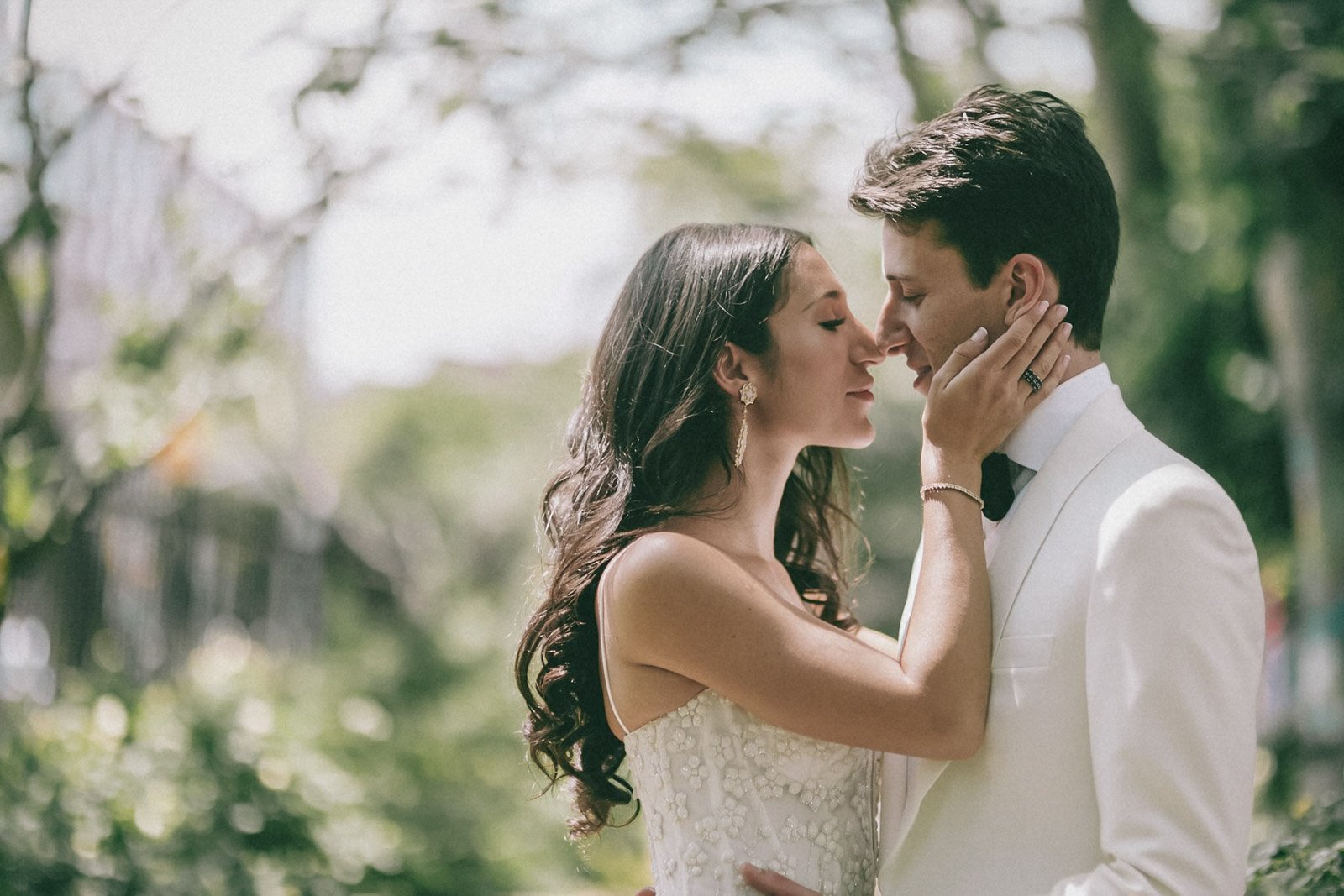 A bride and groom standing closely outdoors, touching foreheads with eyes closed, in a romantic moment surrounded by greenery.
