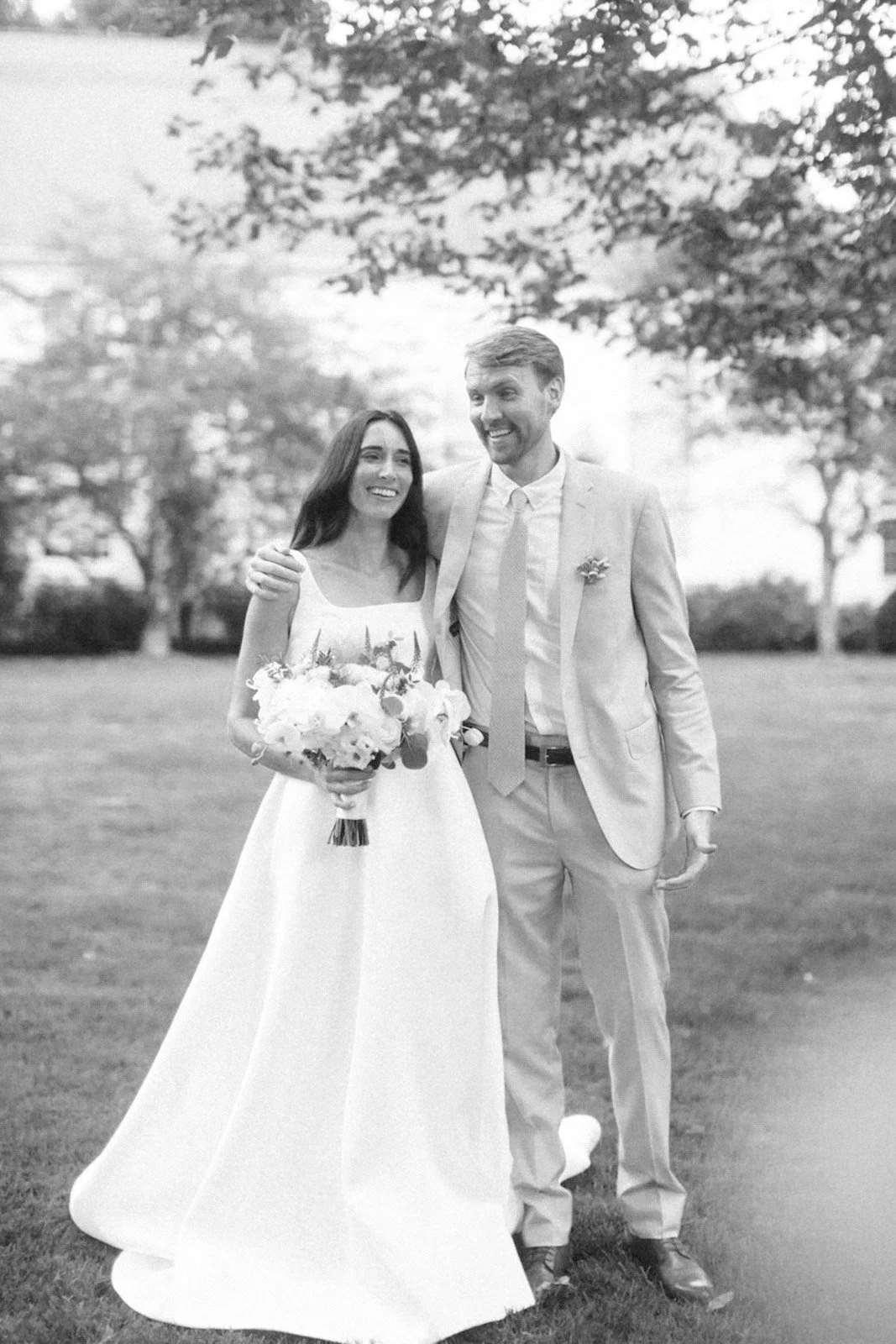 Black and white photo of a newlywed couple walking outdoors on grass, smiling happily. The bride is holding a bouquet of flowers, wearing a sleeveless wedding dress, and the groom is in a light-colored suit with a tie and boutonniere, with his arm ar