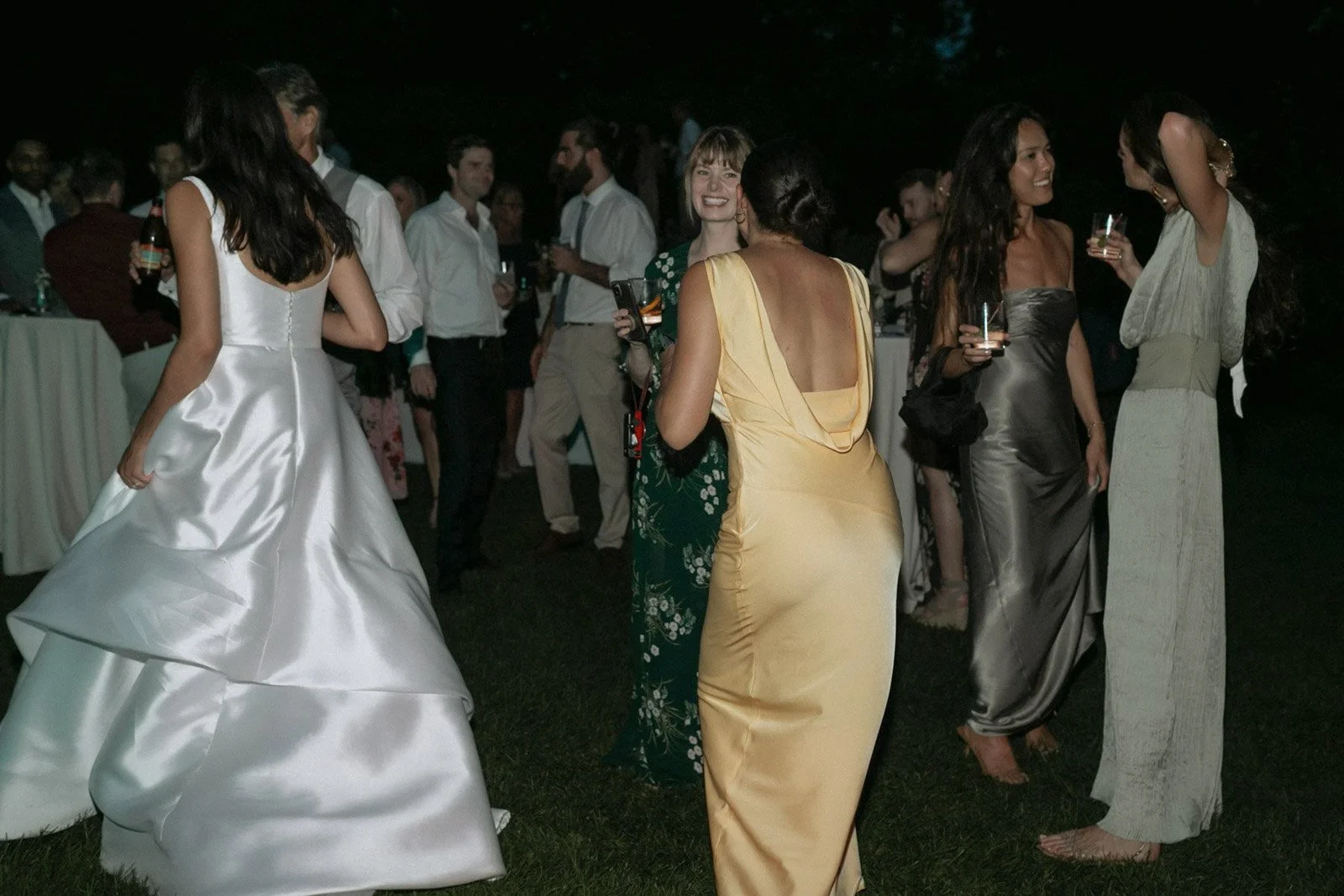 Group of people at an outdoor evening event, dressed in formal attire, socializing and holding drinks.