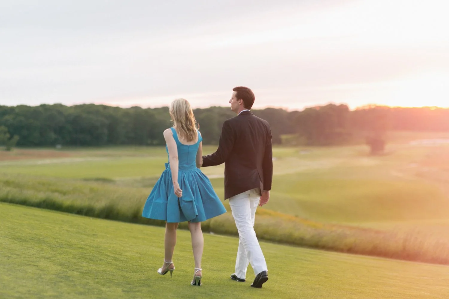 A man and woman walking outdoors on a grassy area during sunset, holding hands. The woman is wearing a blue dress and heels, and the man is dressed in a dark jacket and white pants.