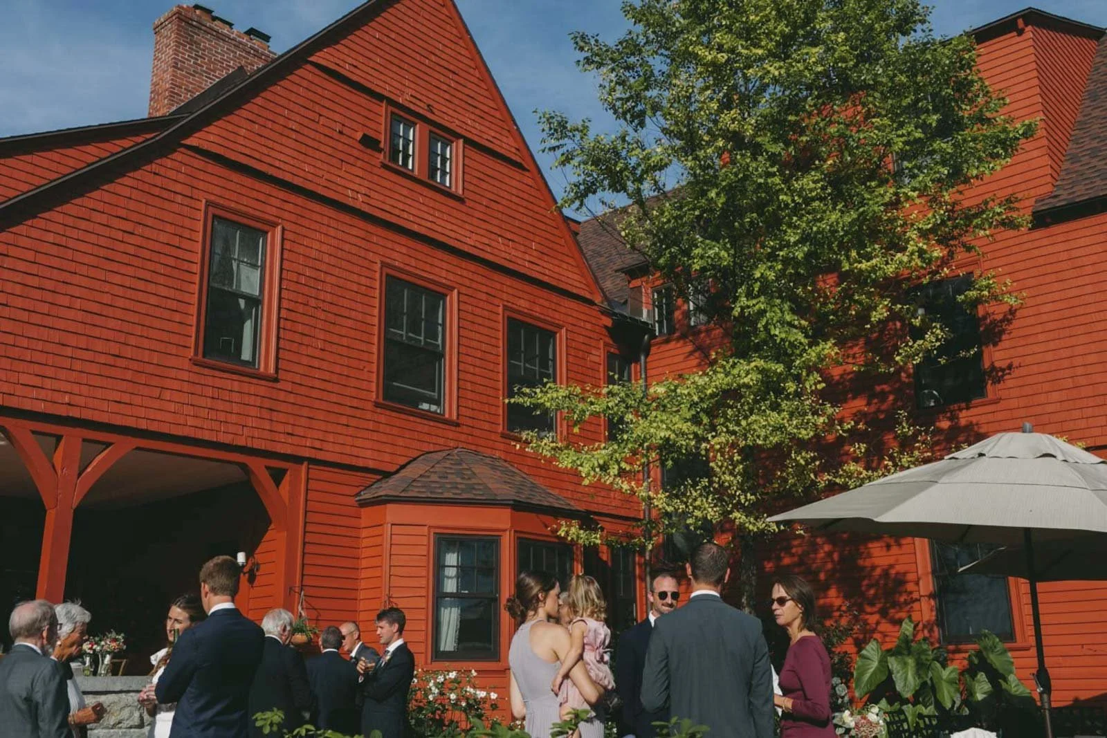 People gathered outside a red wooden house with a tree, some with drinks, during a sunny day.