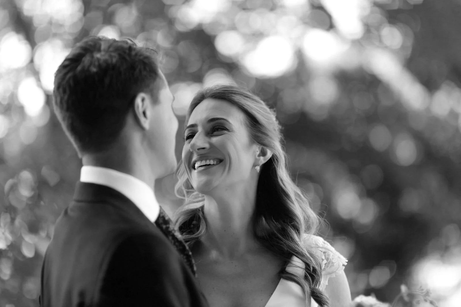 A black and white photo of a bride and groom looking at each other smiling, outdoors with a blurred background of trees.