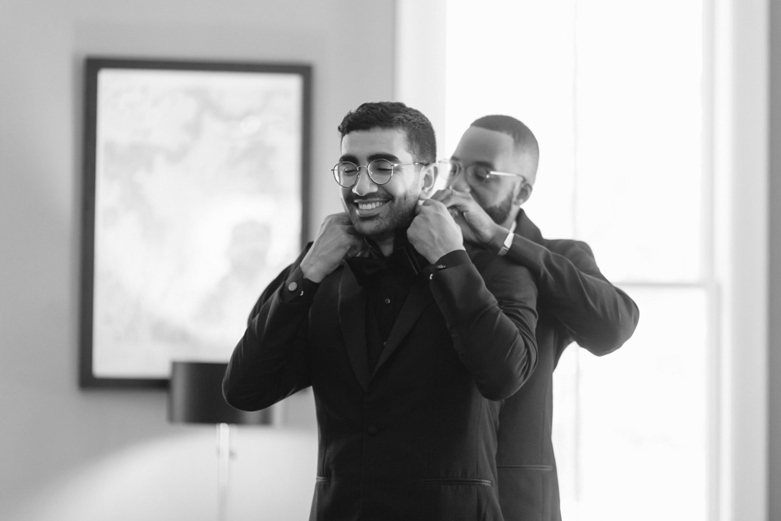 Two men in formal attire, one helping the other adjust his earpiece, smiling in a well-lit room.