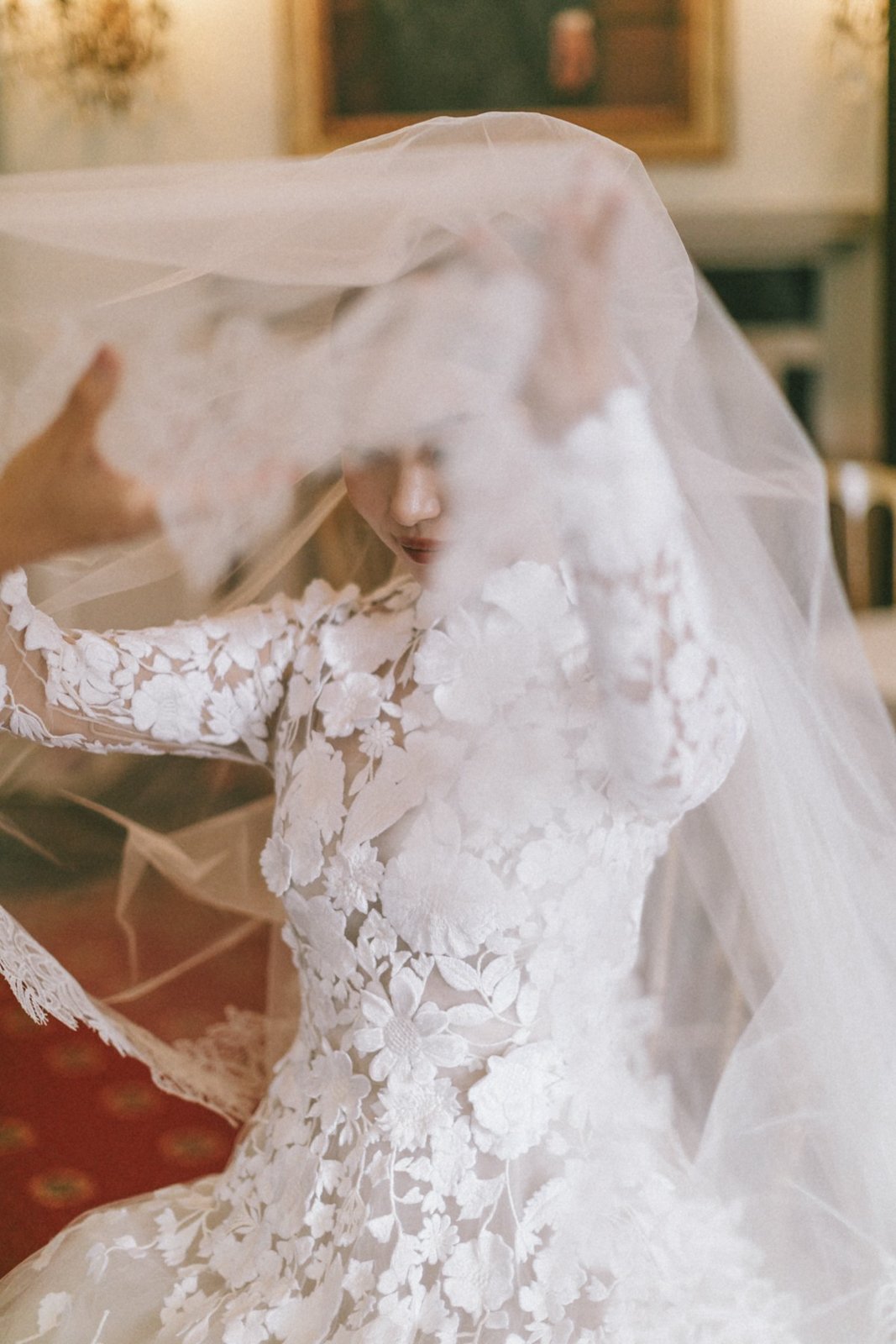 A bride in a lace wedding dress with floral embroidery, partially covered by a sheer veil, as someone adjusts or drapes the veil over her head.
