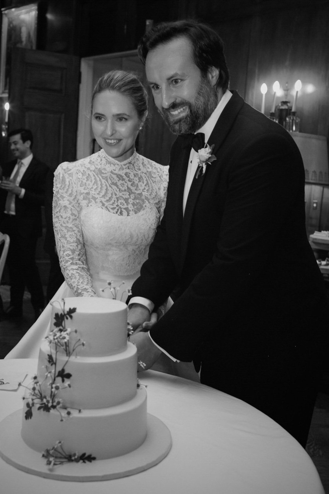 Black and white photo of a bride and groom cutting a wedding cake at their wedding reception, with guests in the background.