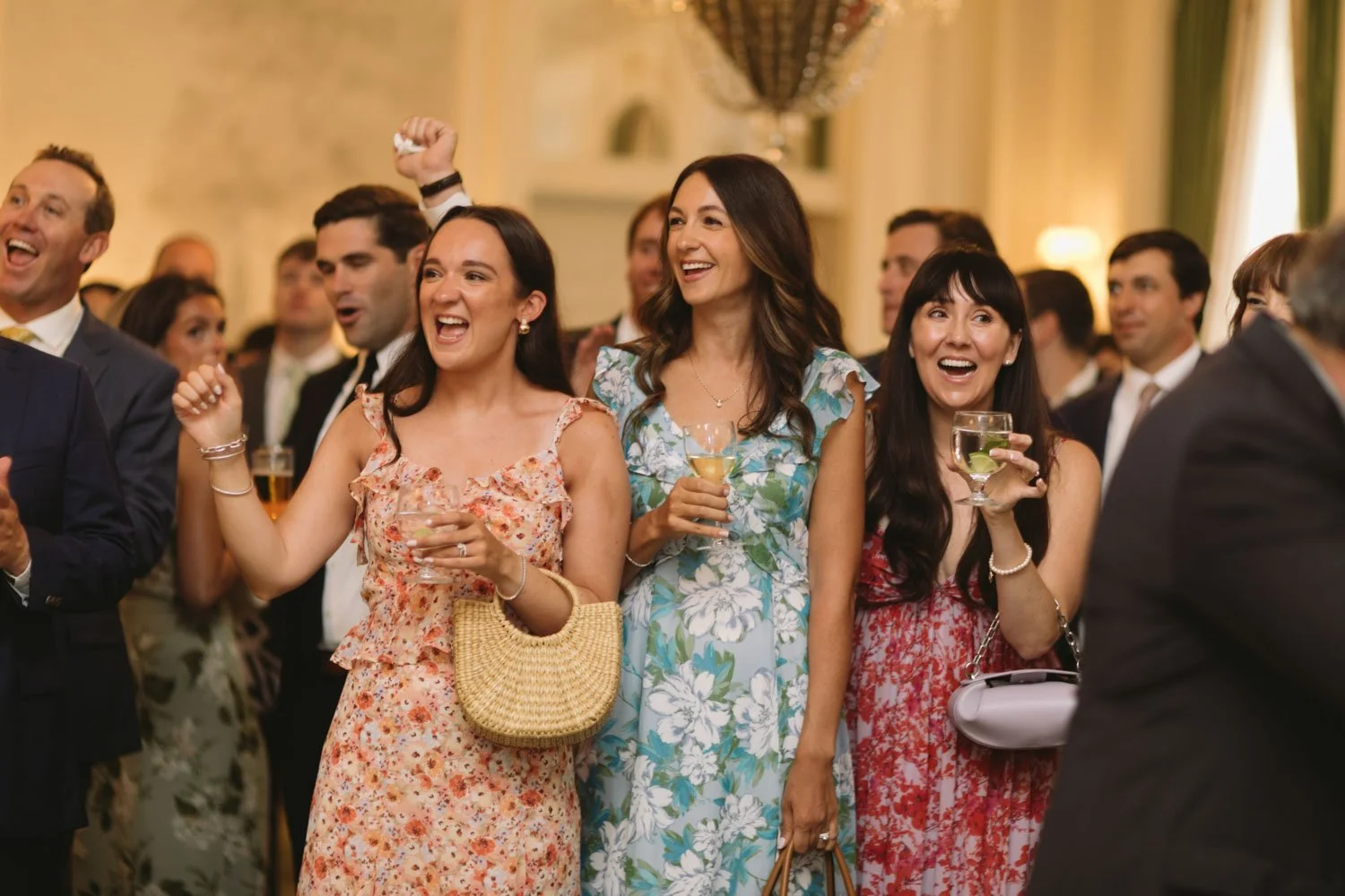 Group of people at a wedding reception, smiling, holding drinks, dressed in formal summer attire, in an elegant indoor setting.