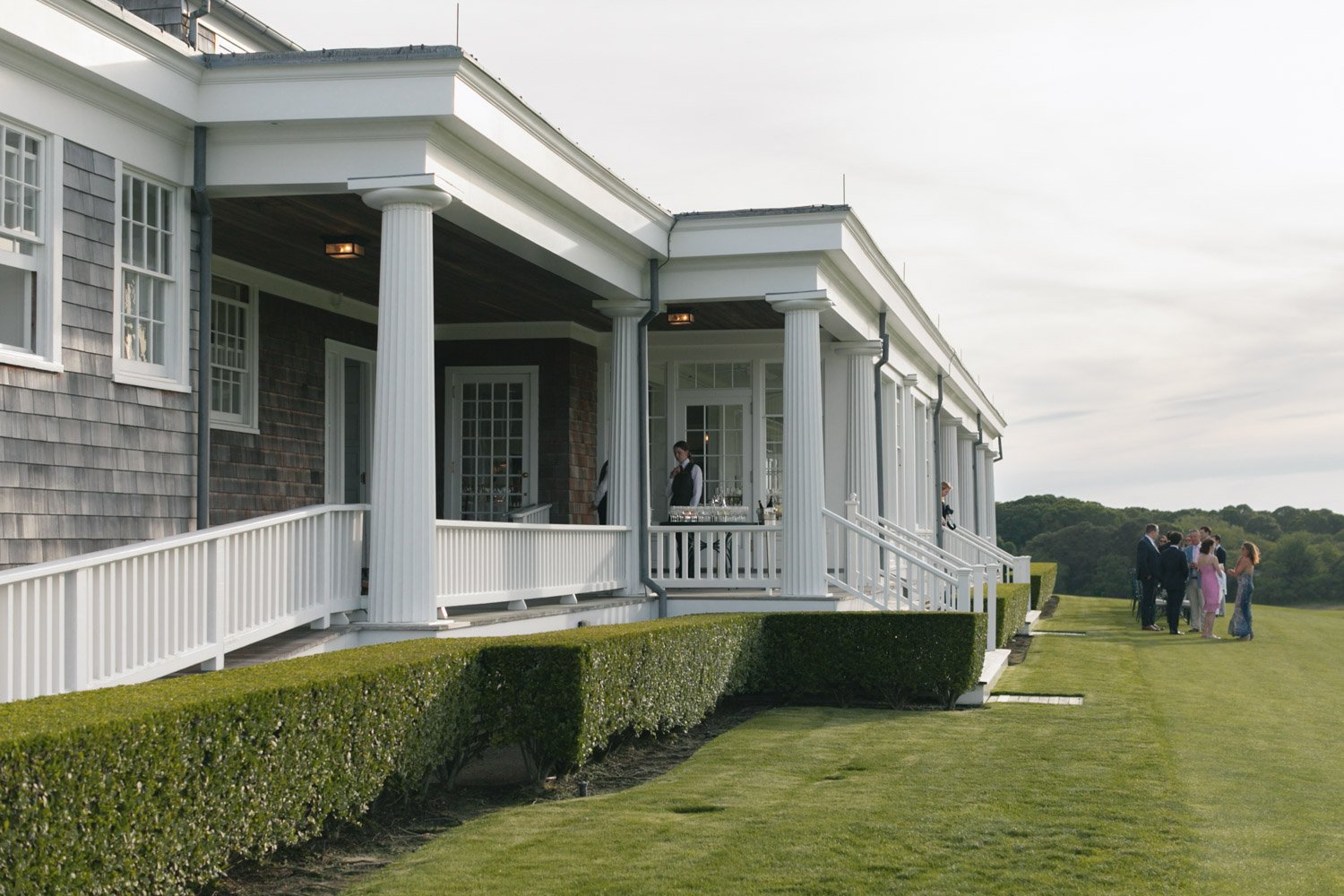 A group of people gathering outside a large, white, colonial-style house with tall columns and a porch. The house is surrounded by well-manicured grass and hedges, with trees in the background, under a partly cloudy sky.