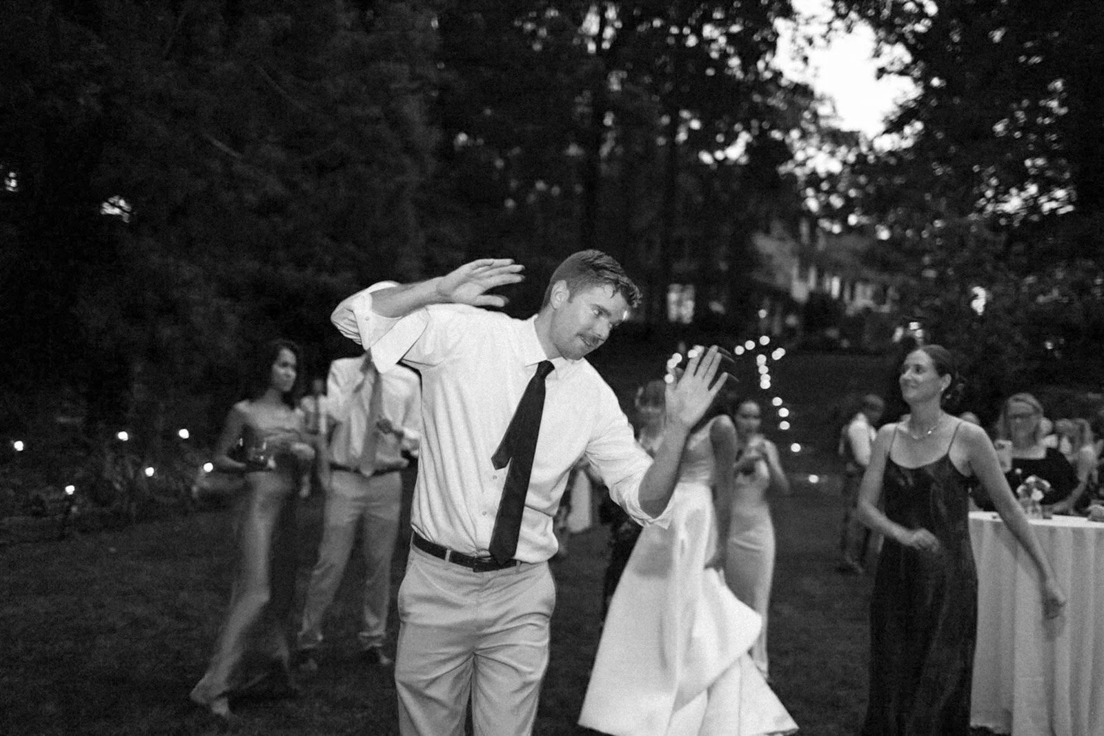 A man dancing at an outdoor evening event, wearing a white shirt and black tie, with an audience and string lights in the background.