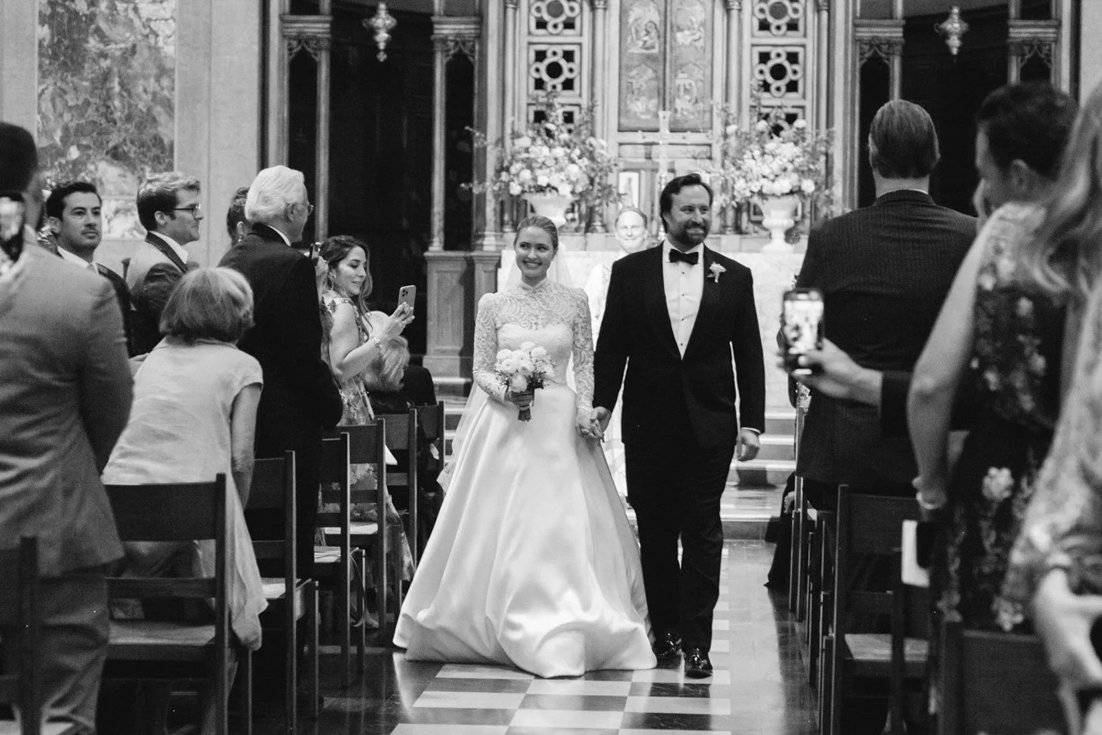 Black and white photo of a wedding ceremony inside a church. The bride and groom are walking down the aisle, holding hands. The bride wears a lace wedding dress and holds a bouquet. The groom wears a tuxedo with a bow tie. Guests are standing on eith