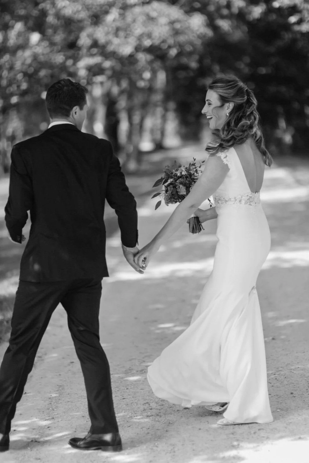 A bride and groom holding hands and walking outdoors, with the bride smiling and holding a bouquet of flowers.