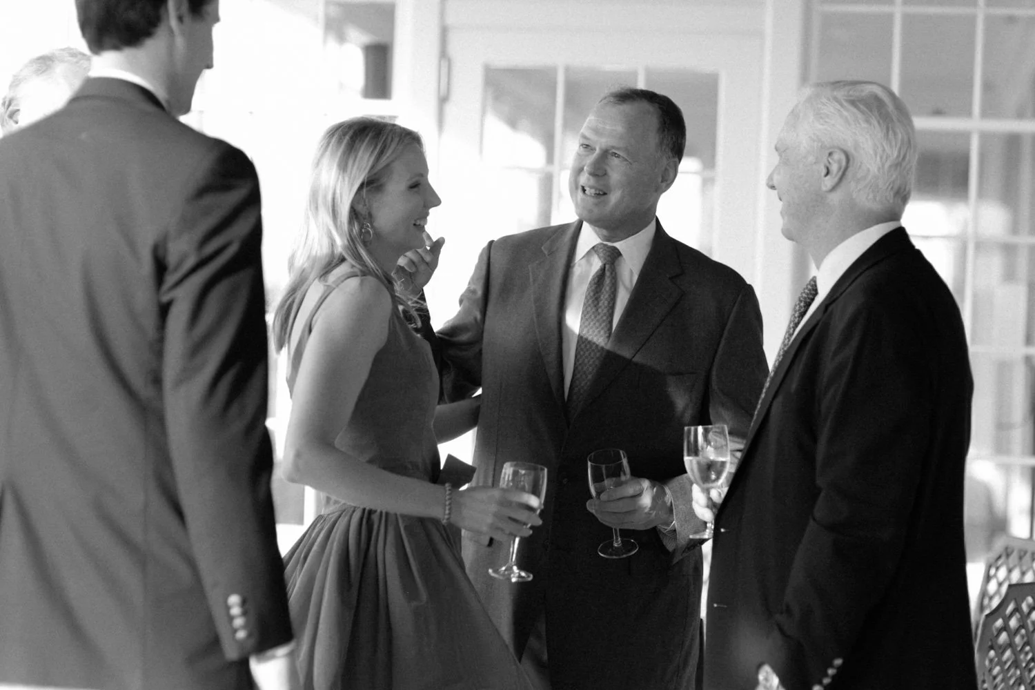 A group of five people, three men and two women, engaged in conversation at a social event, holding wine glasses, dressed in formal attire, in a well-lit indoor setting.