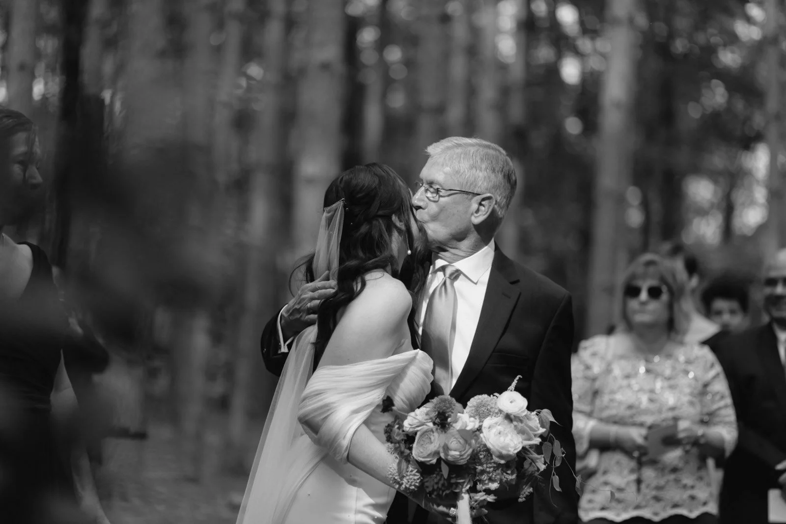 An elderly man in a suit kisses a woman in a wedding gown on the cheek during a wedding ceremony outdoors, with friends and family in the background.