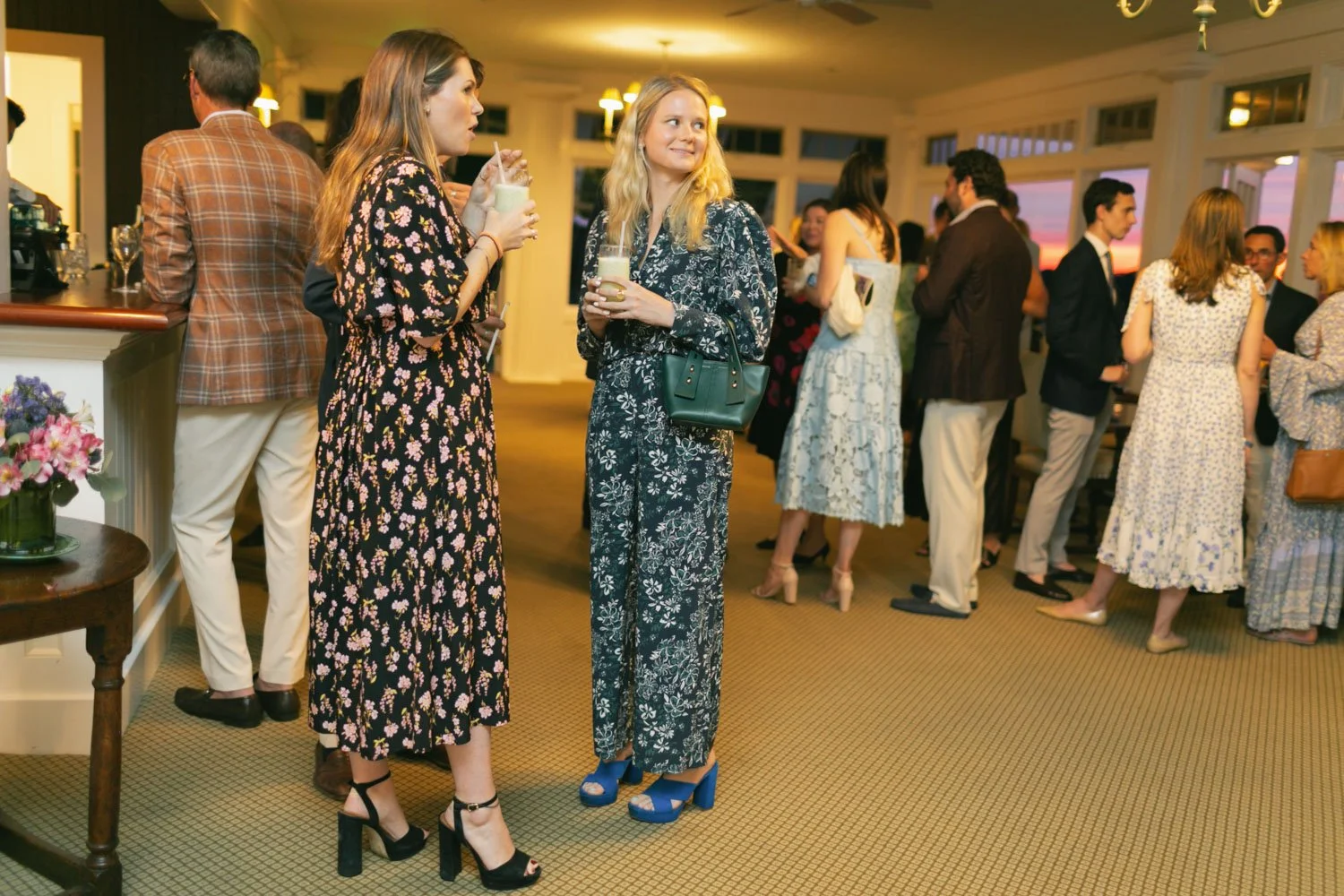People socializing at an indoor event in the evening, some holding drinks and engaged in conversation, dressed in semi-formal and fashionable clothing.