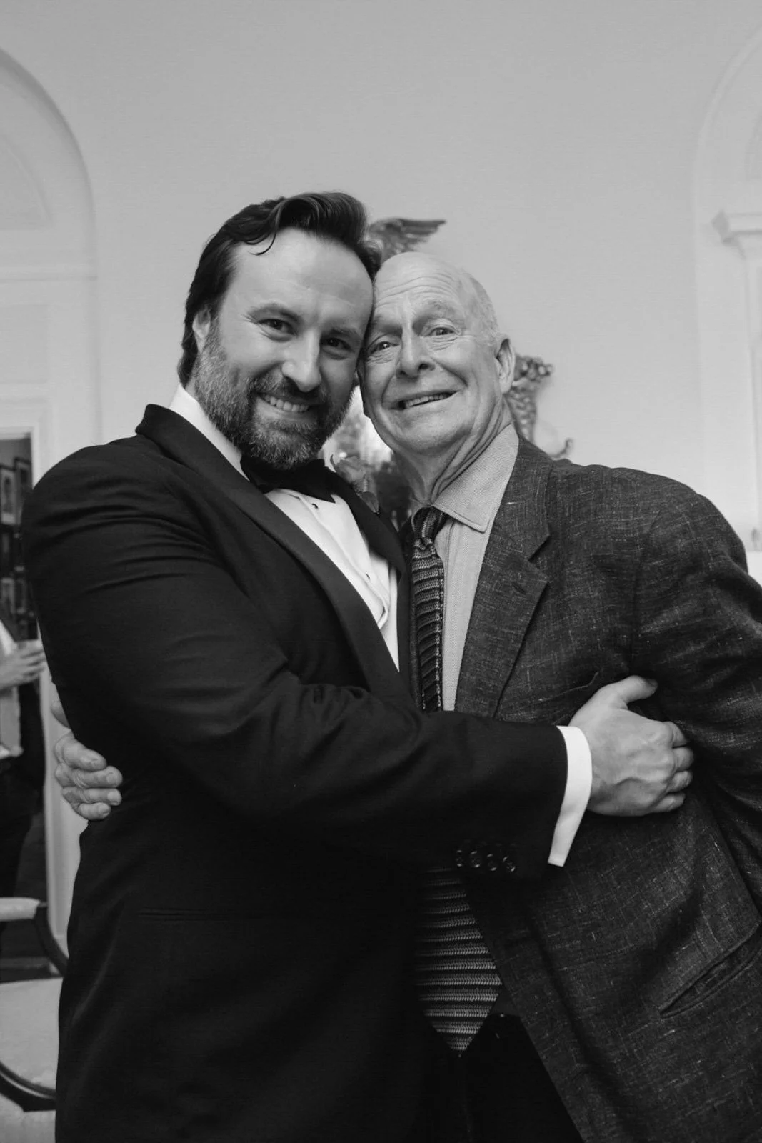 Two men embracing and smiling at a formal event in a room with white walls and arched doorways.