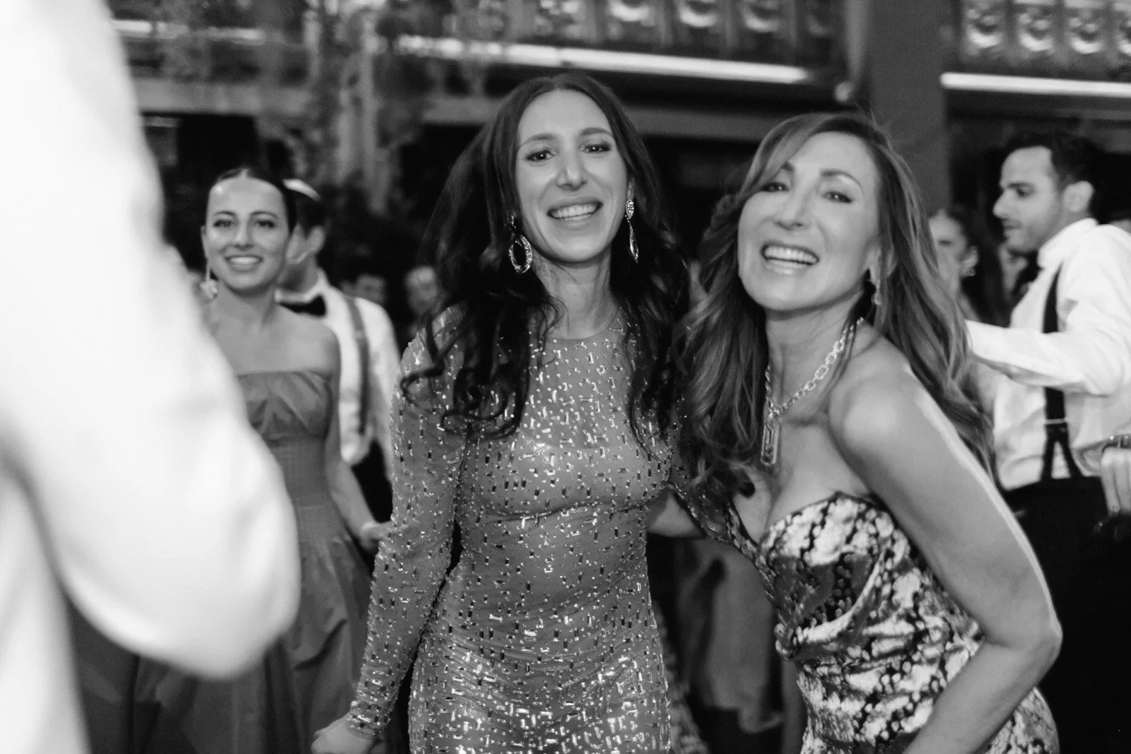 Black and white photo of women at party or event, two women in center smiling and posing for camera, wearing elegant dresses, woman on left has long dark hair and earrings, woman on right has shoulder-length light hair and necklace, other people visi