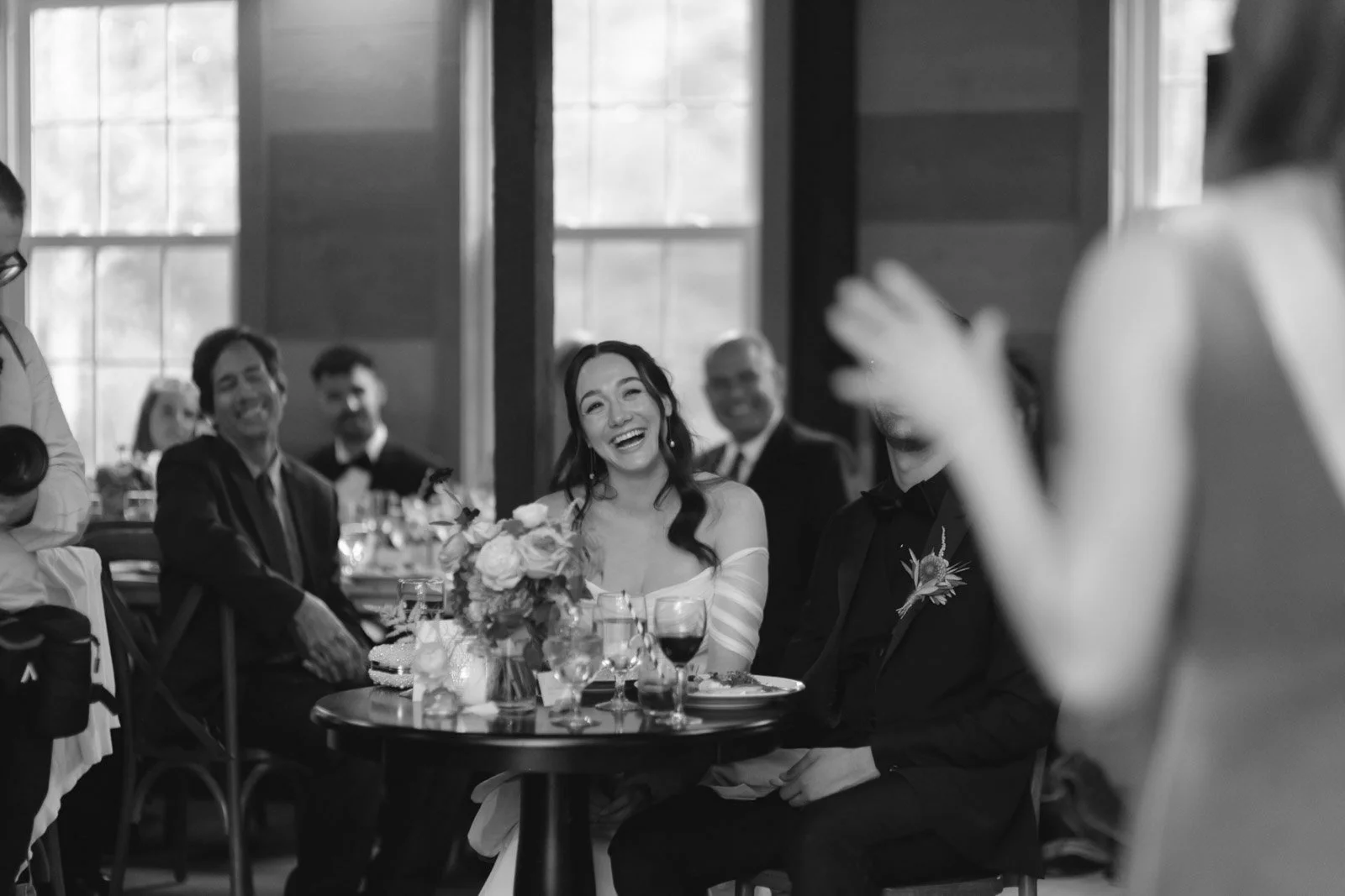 A black and white photo of a joyful woman in a wedding dress holding a bouquet, seated at a table with friends and family, some are smiling and laughing, at what appears to be a wedding reception.
