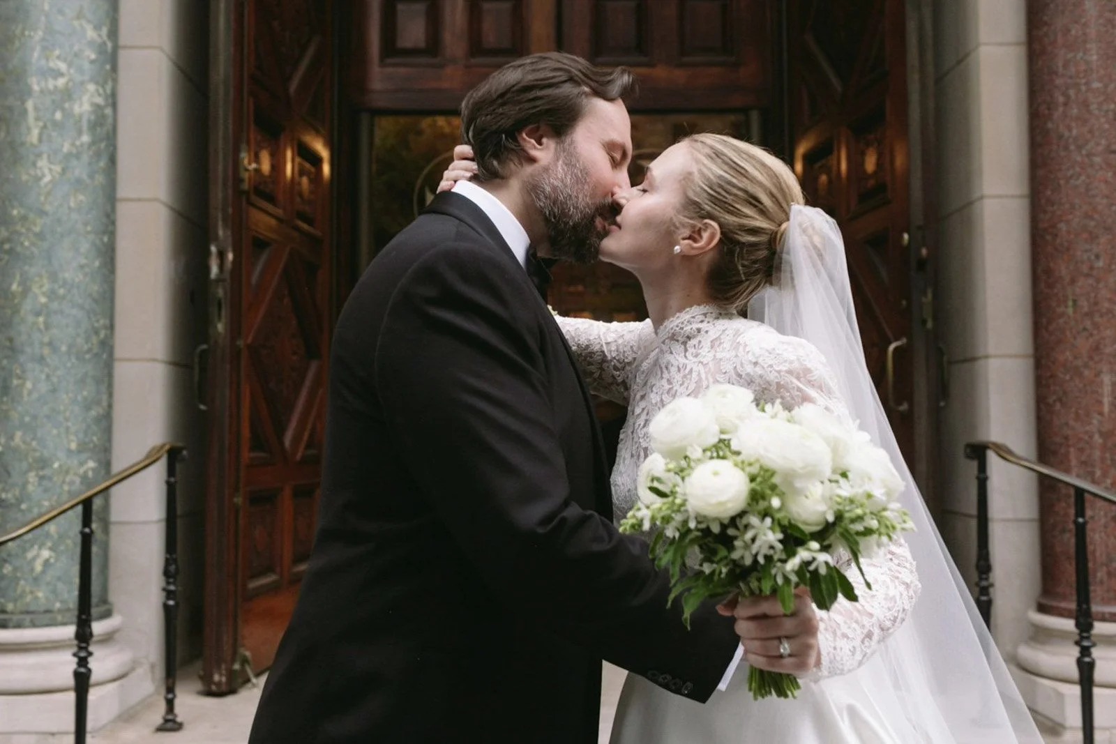 A newlywed couple sharing a kiss outside a building with wooden doors, the bride holding a bouquet of white flowers and wearing a lace wedding dress with a veil, the groom in a black tuxedo.