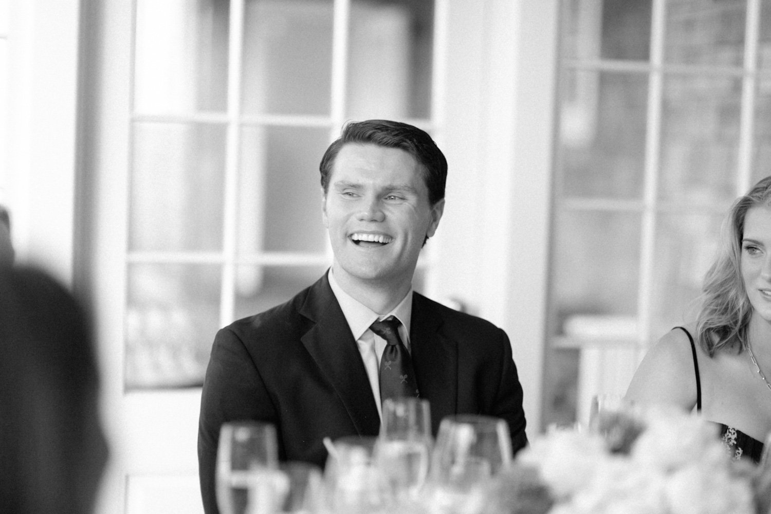 Black and white photo of a man in a tuxedo smiling at an indoor social gathering, with a woman partially visible beside him and glasses on the table in the foreground.