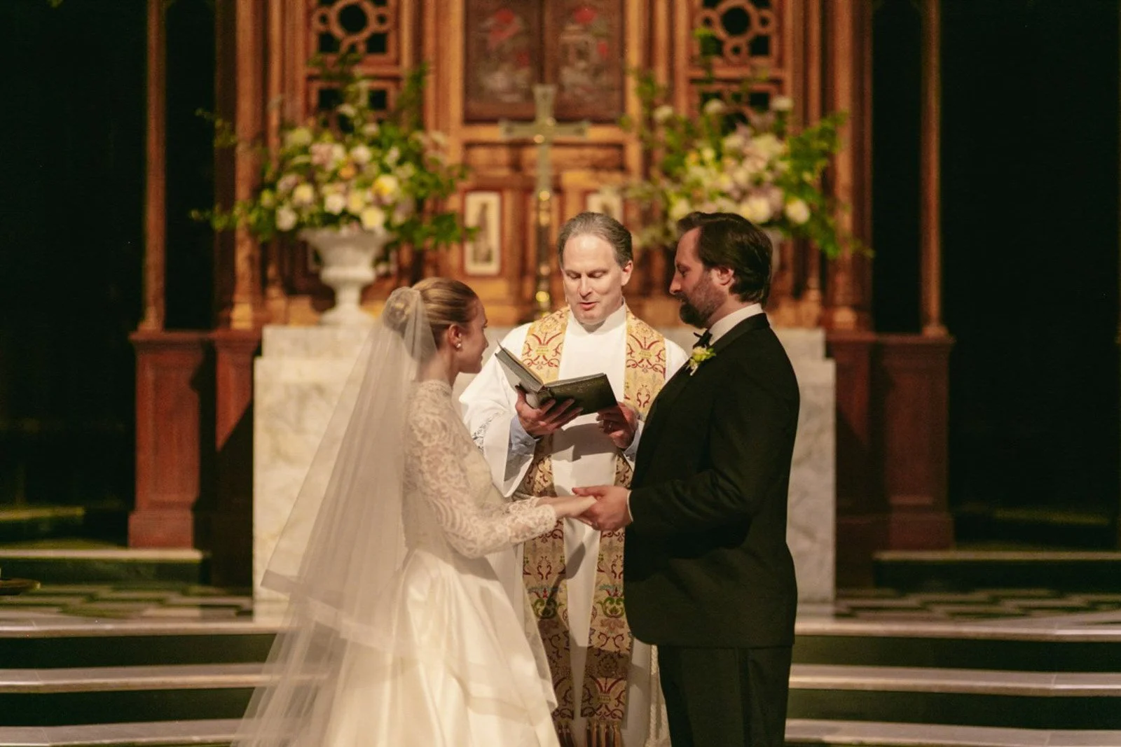 A wedding ceremony inside a church with a bride and groom holding hands in front of a priest. The bride is wearing a white lace wedding dress and veil, while the groom is in a black tuxedo. The priest is in religious robes and holding an open book. T