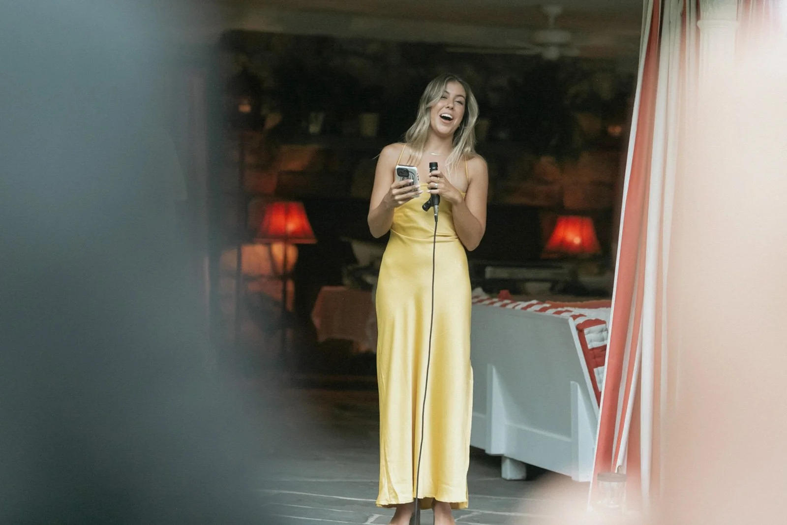 A young woman in a long yellow dress is singing or speaking into a microphone in a cozy indoor setting with warm lighting and red lamps in the background.