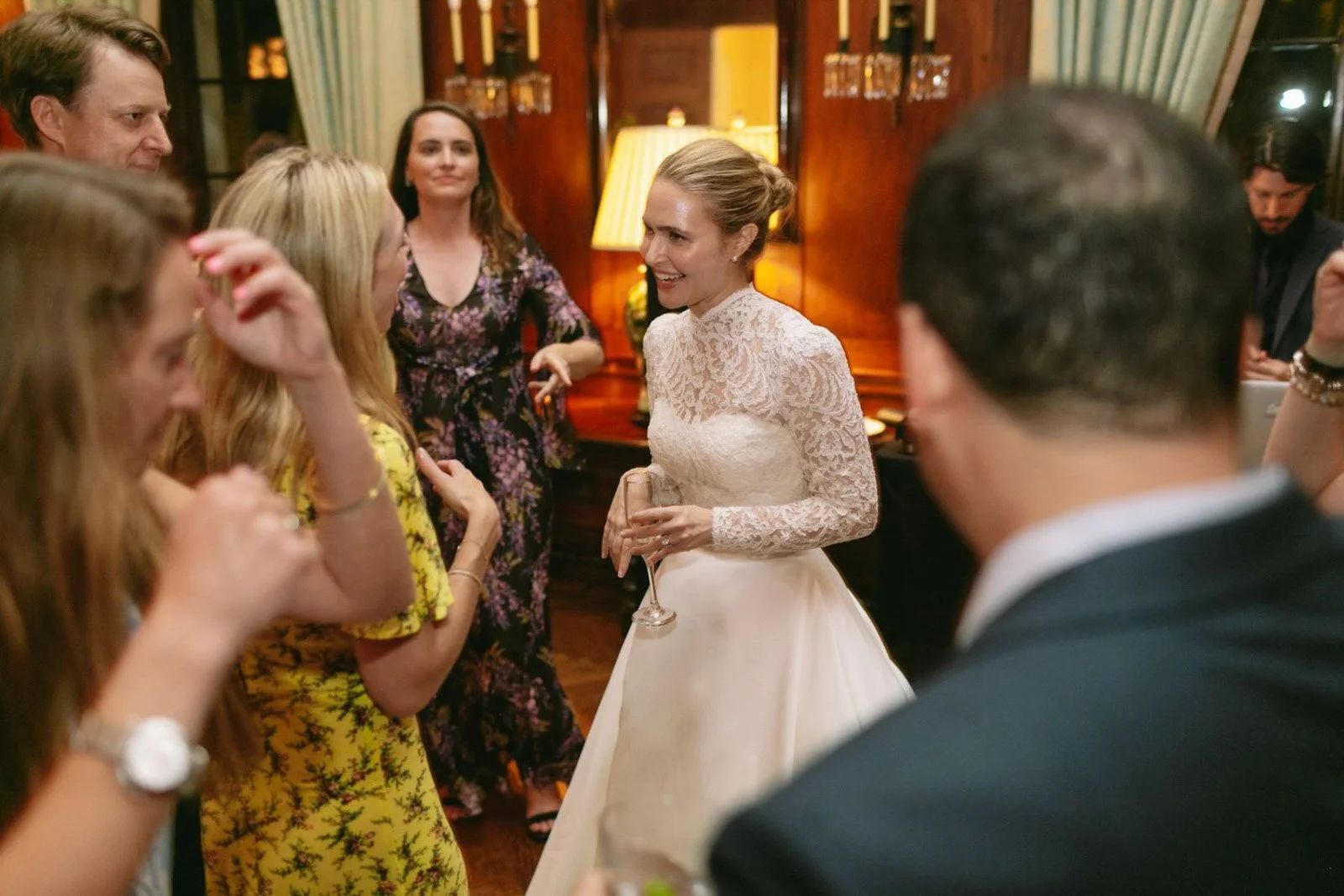 A bride in a lace wedding dress talking to guests at her reception, holding a glass of wine, surrounded by friends and family in a warmly lit room.