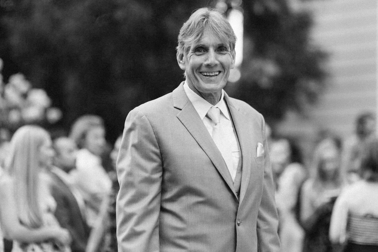 A smiling man in a suit and tie standing outdoors at an event, with a group of people blurred in the background.