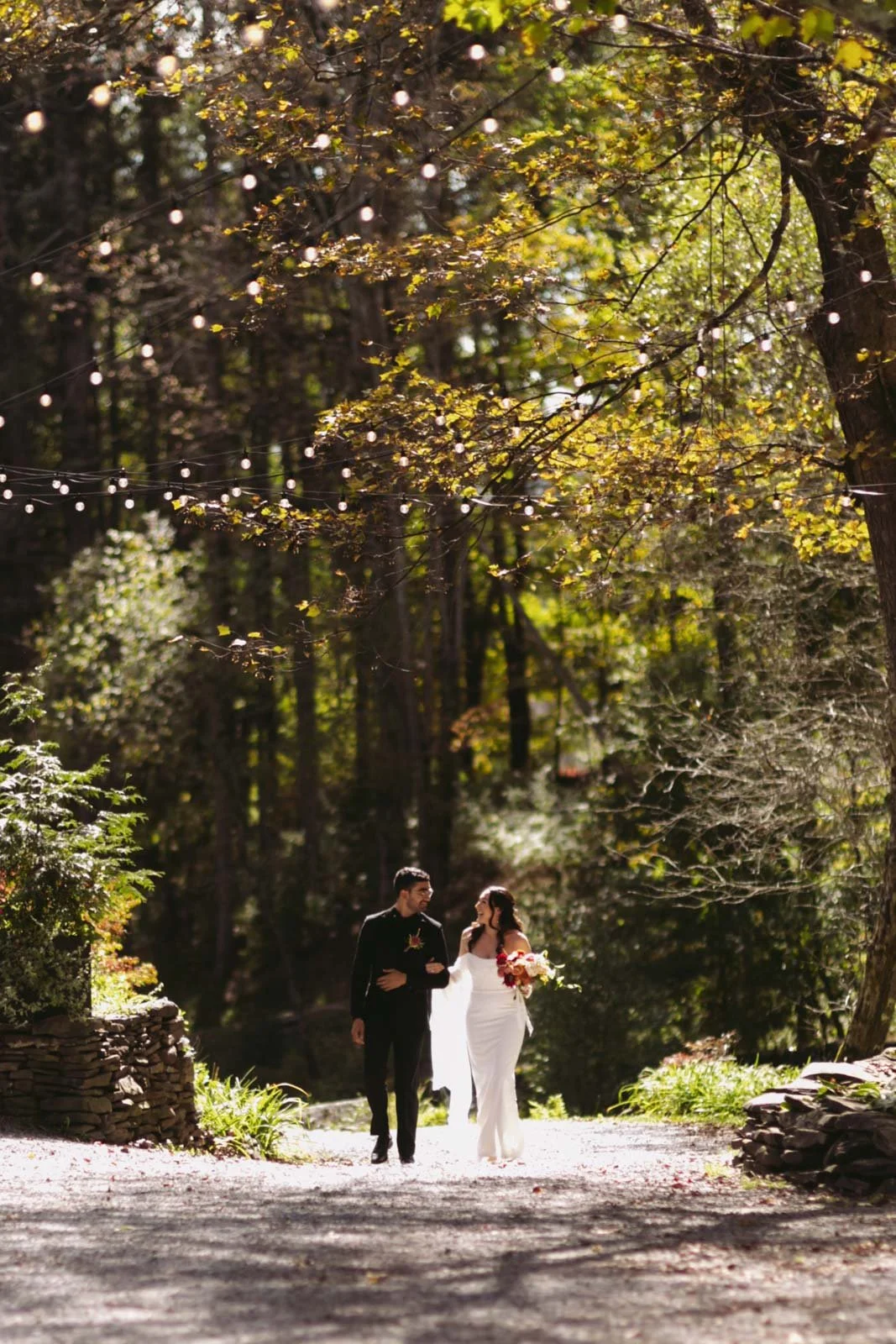 A bride and groom walk together through a sunlit wooded outdoor setting, with the bride holding a bouquet and the groom wearing a black suit. String lights hang overhead amidst the trees.