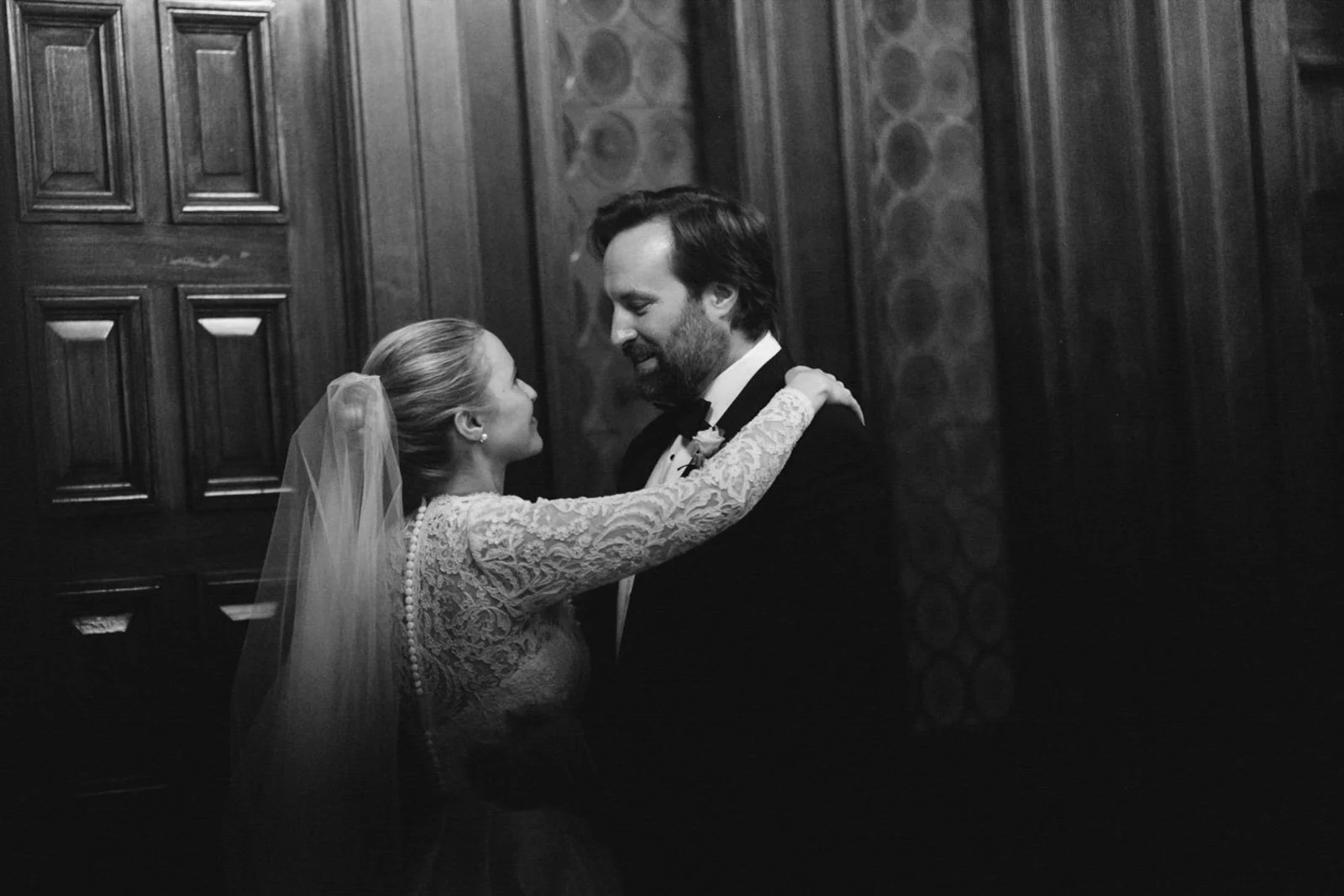 A bride and groom share a dance during their wedding reception in a wood-paneled room, smiling at each other.