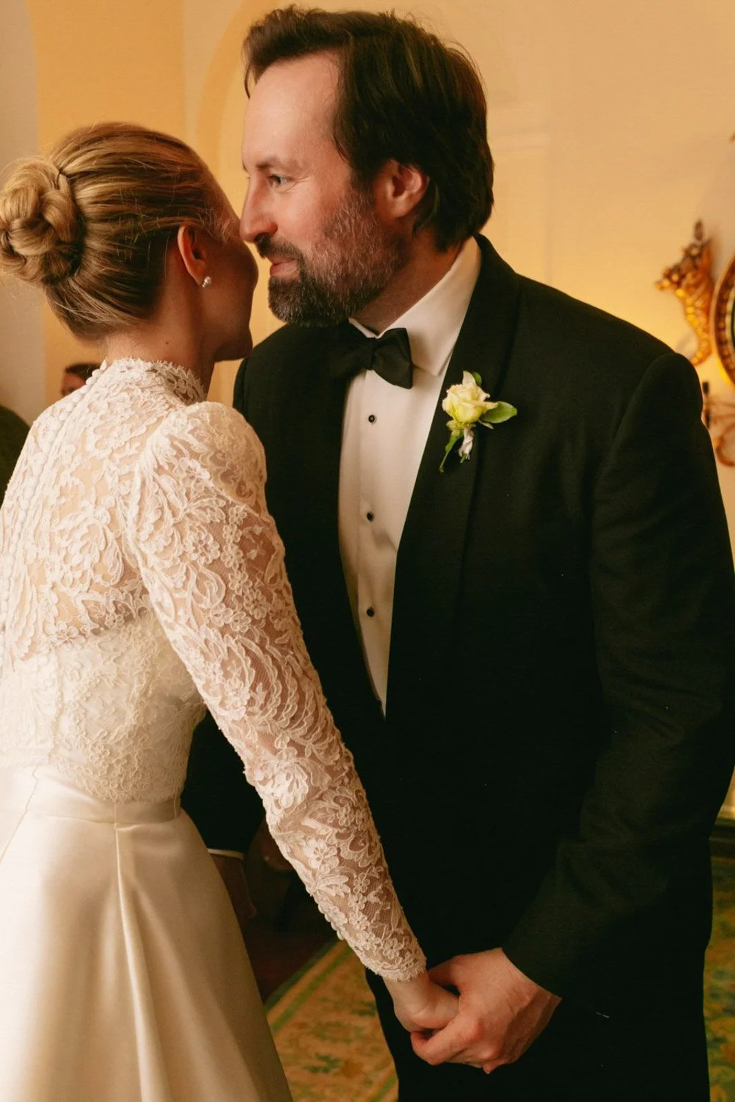 A bride and groom holding hands during their wedding, standing close with foreheads touching, inside a warmly lit room.