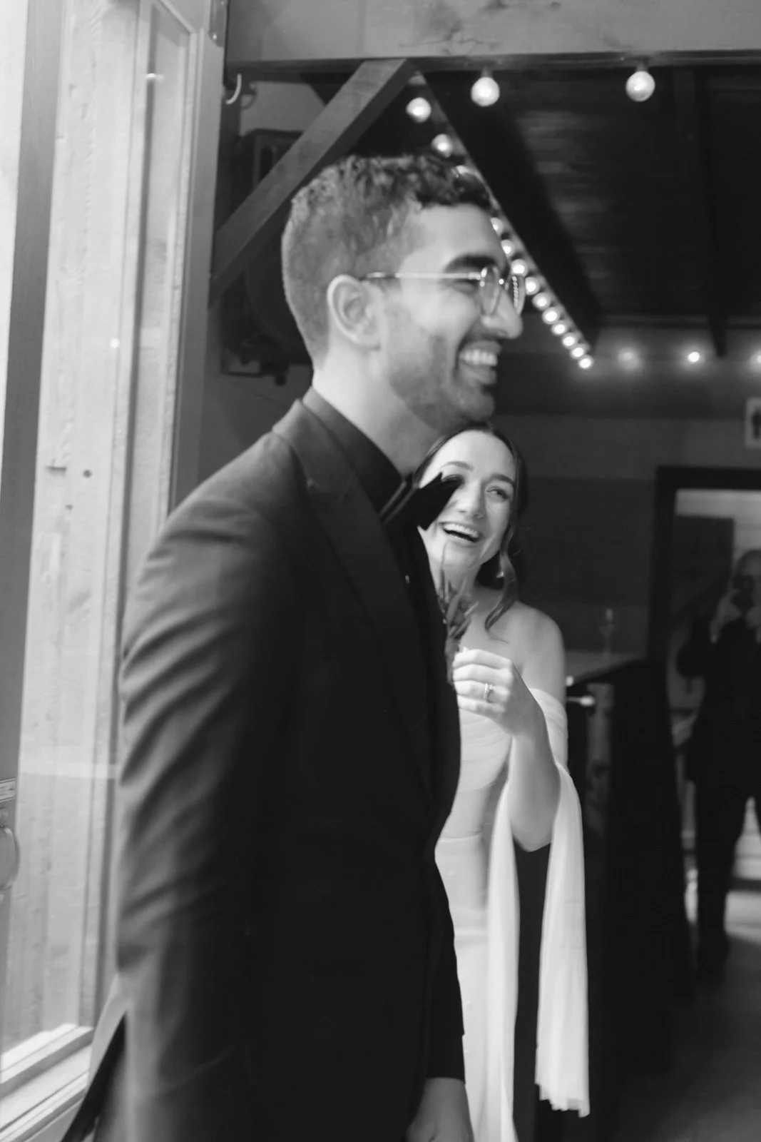 A black and white photo of a man in a suit with glasses, smiling, with a woman in an off-shoulder dress laughing behind him. They appear to be at a celebration or formal event, with string lights overhead.