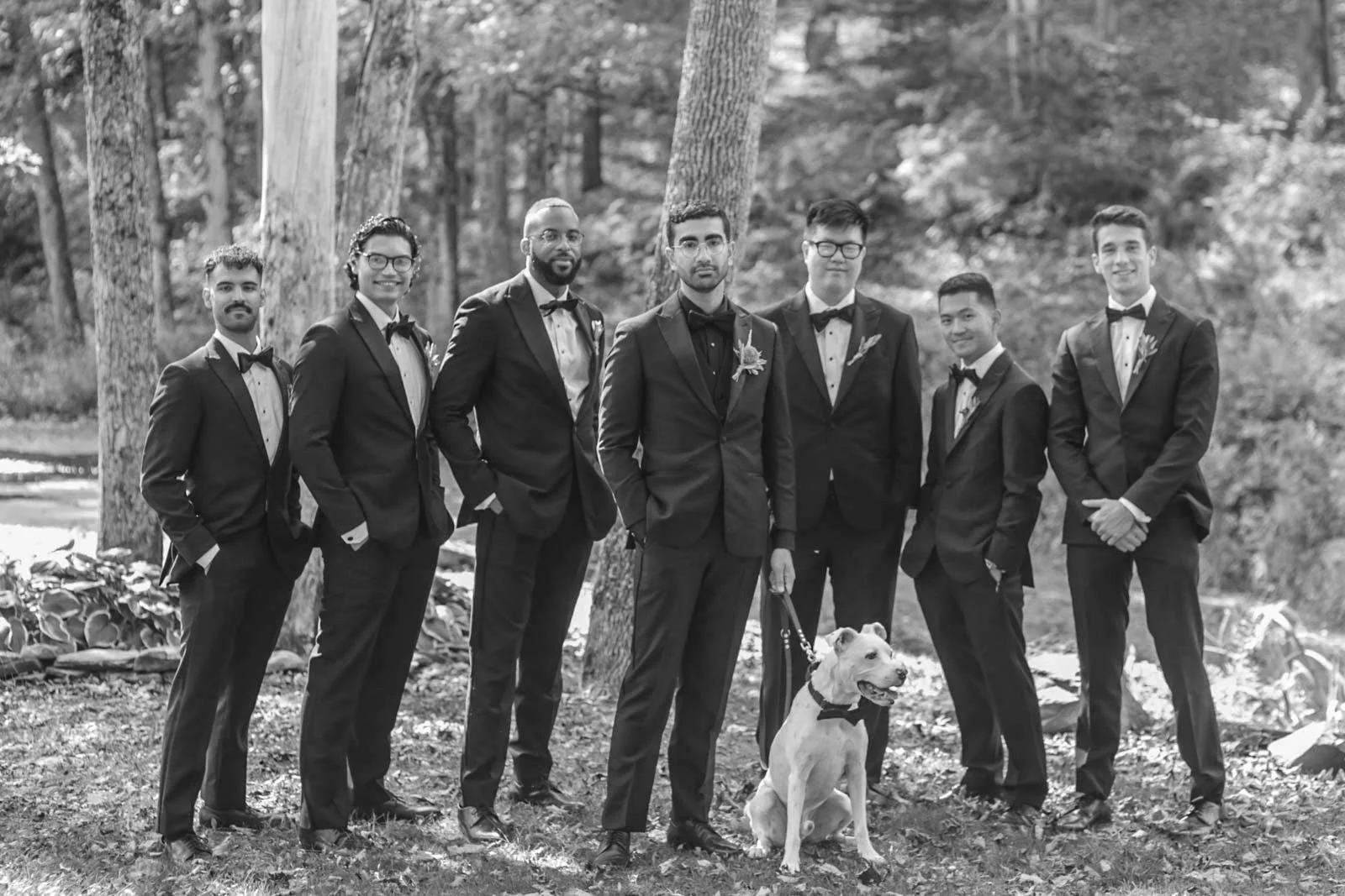 A group of seven men in black tuxedos with bow ties, standing outdoors on grass among trees, with a dog sitting in front of them, all looking at the camera, during daytime in a park or forest setting.