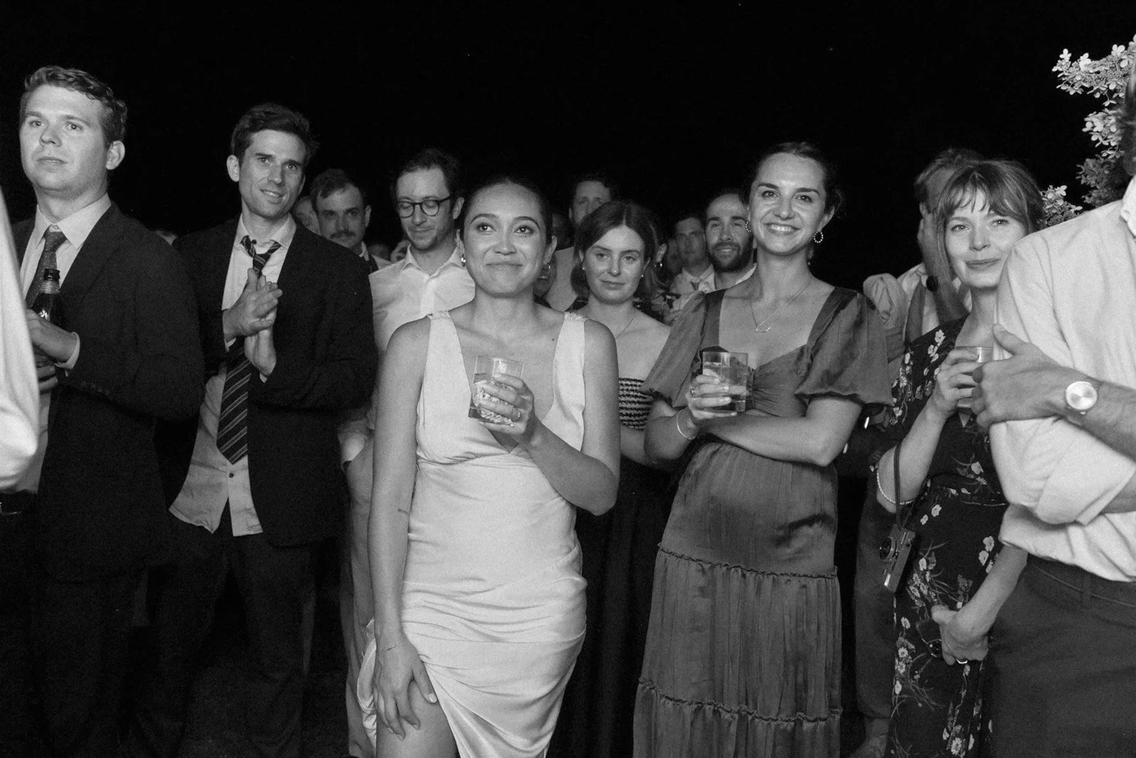 Group of people at a social event, some holding drinks, smiling and mingling outdoors at night.