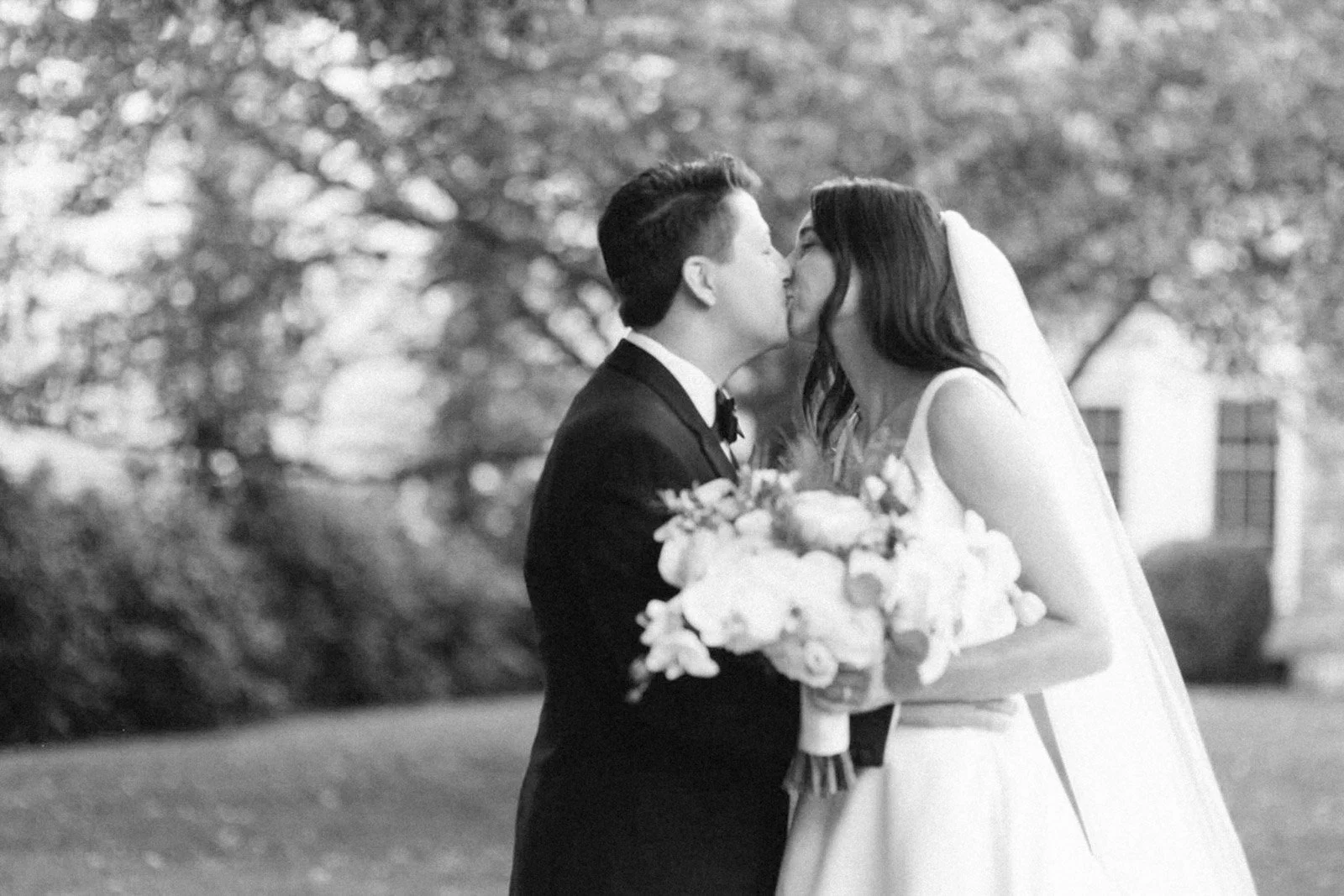 A black and white photo of a couple in wedding attire sharing a kiss outdoors, with trees and a building in the background.