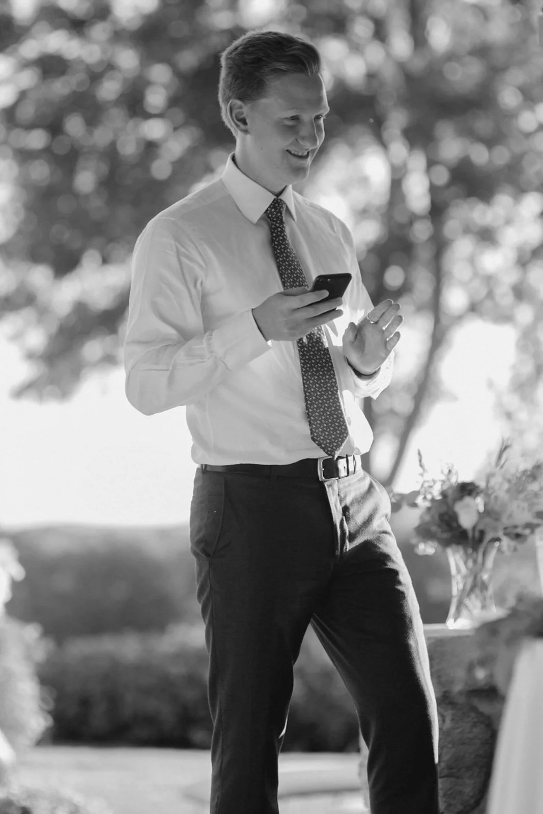 A young man dressed in a white shirt, patterned tie, and dark trousers is outdoors, smiling and looking at his smartphone.