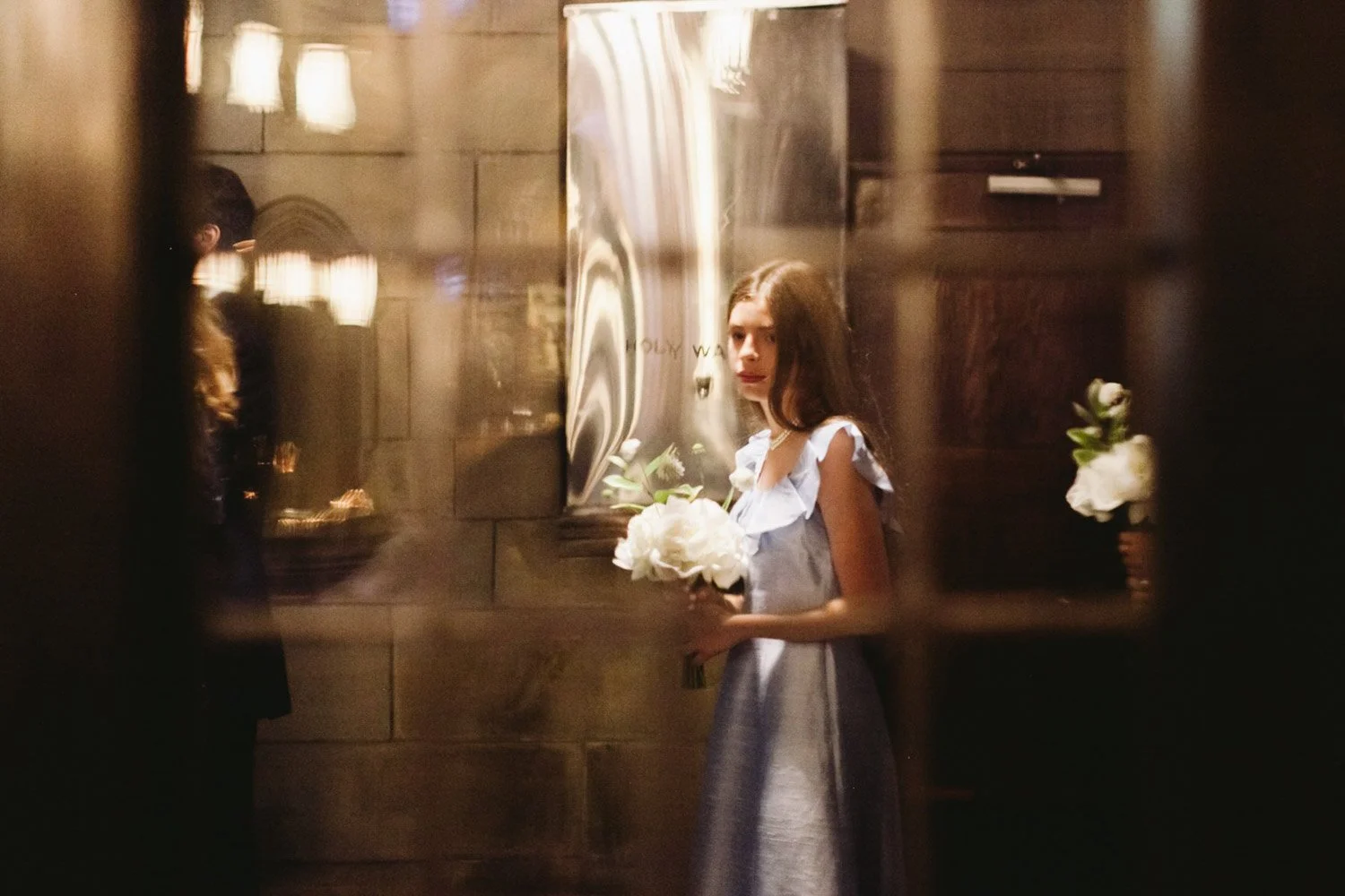 A young woman in a light blue dress holding a bouquet of white flowers, looking through a glass window or mirror in a dimly lit indoor setting.