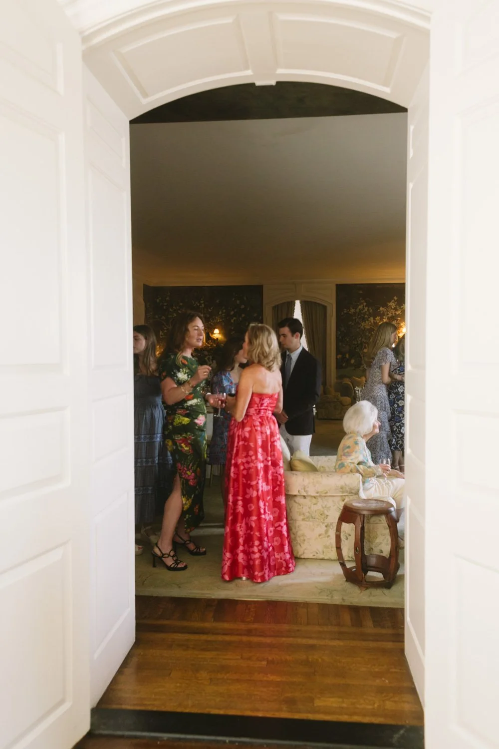 People socializing at an indoor gathering, viewed through a doorframe, with women wearing floral and elegant dresses and men in suits.