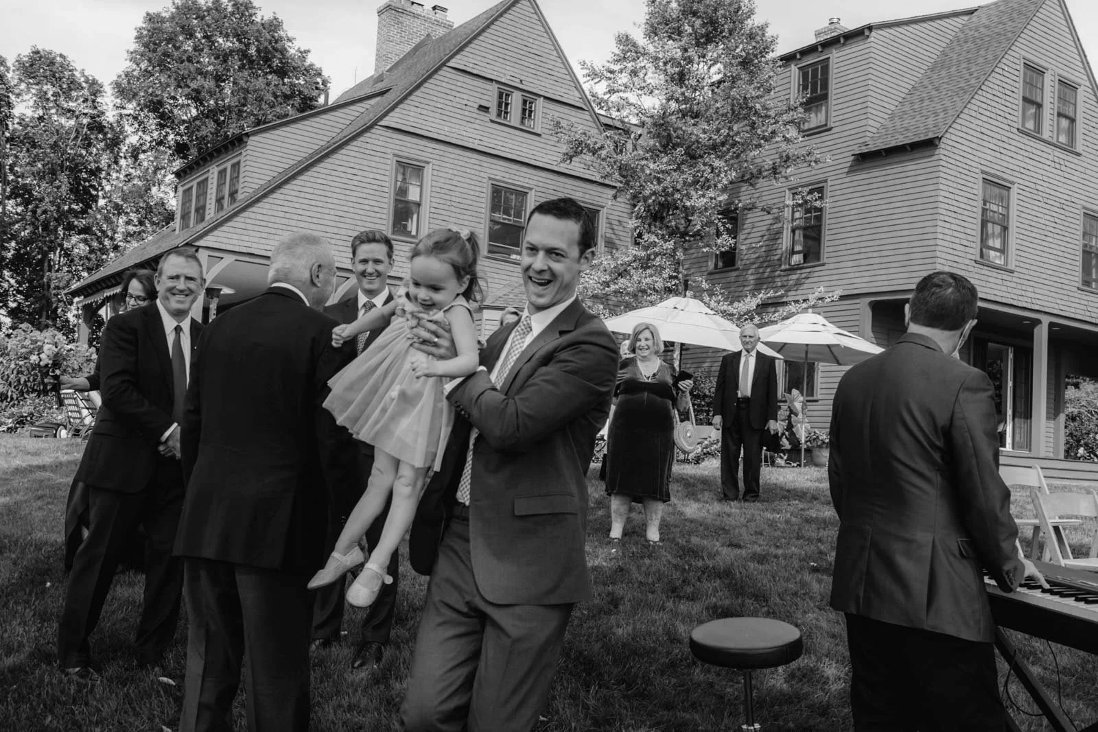 A joyful outdoor gathering of people dressed formally, some in suits and dresses, with a large house in the background. One man holds a young girl who is smiling, and a person in the background is playing a keyboard. Some guests are standing under um