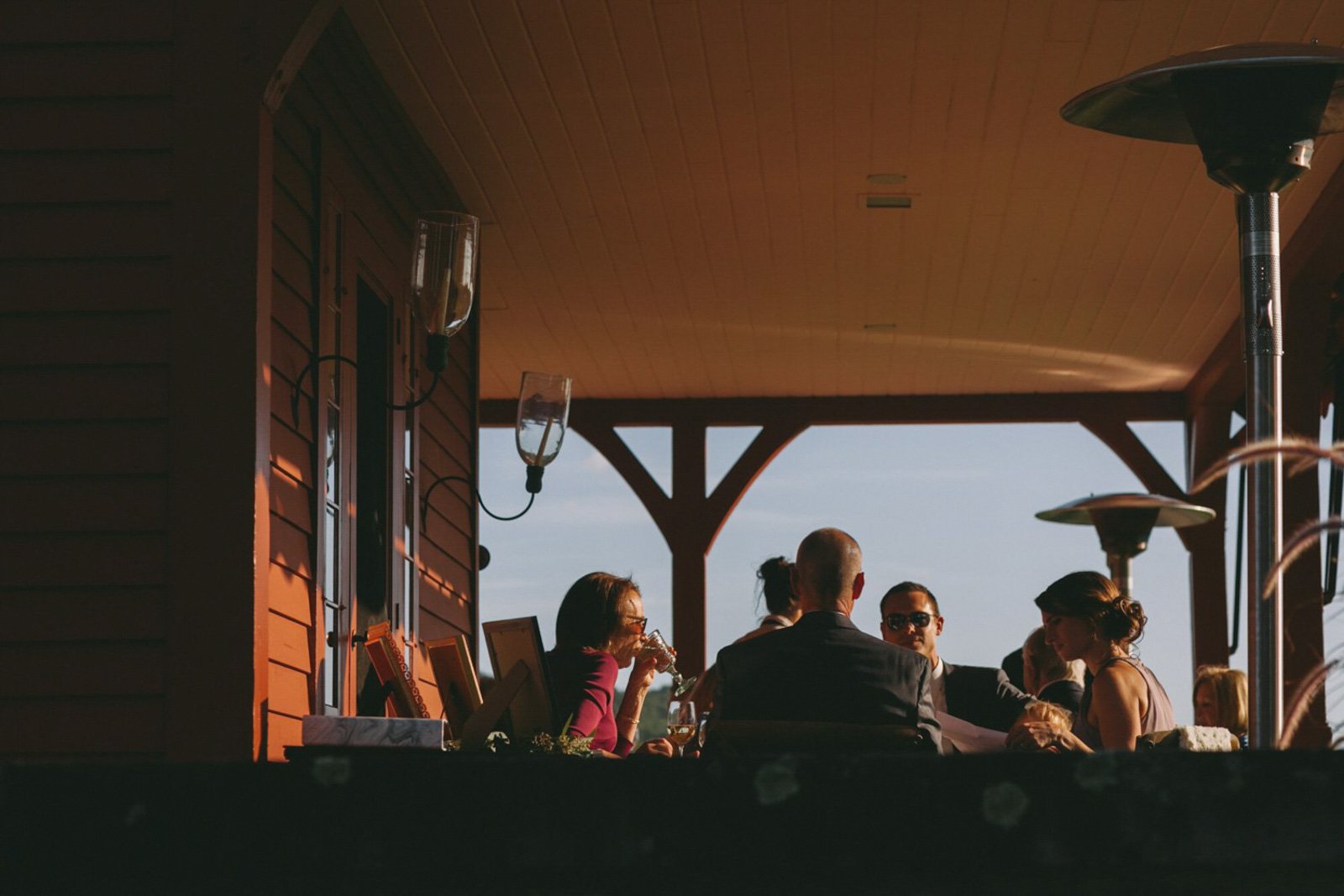 People sitting at a table on an outdoor patio, enjoying drinks and conversation, with a cloudy sky in the background.
