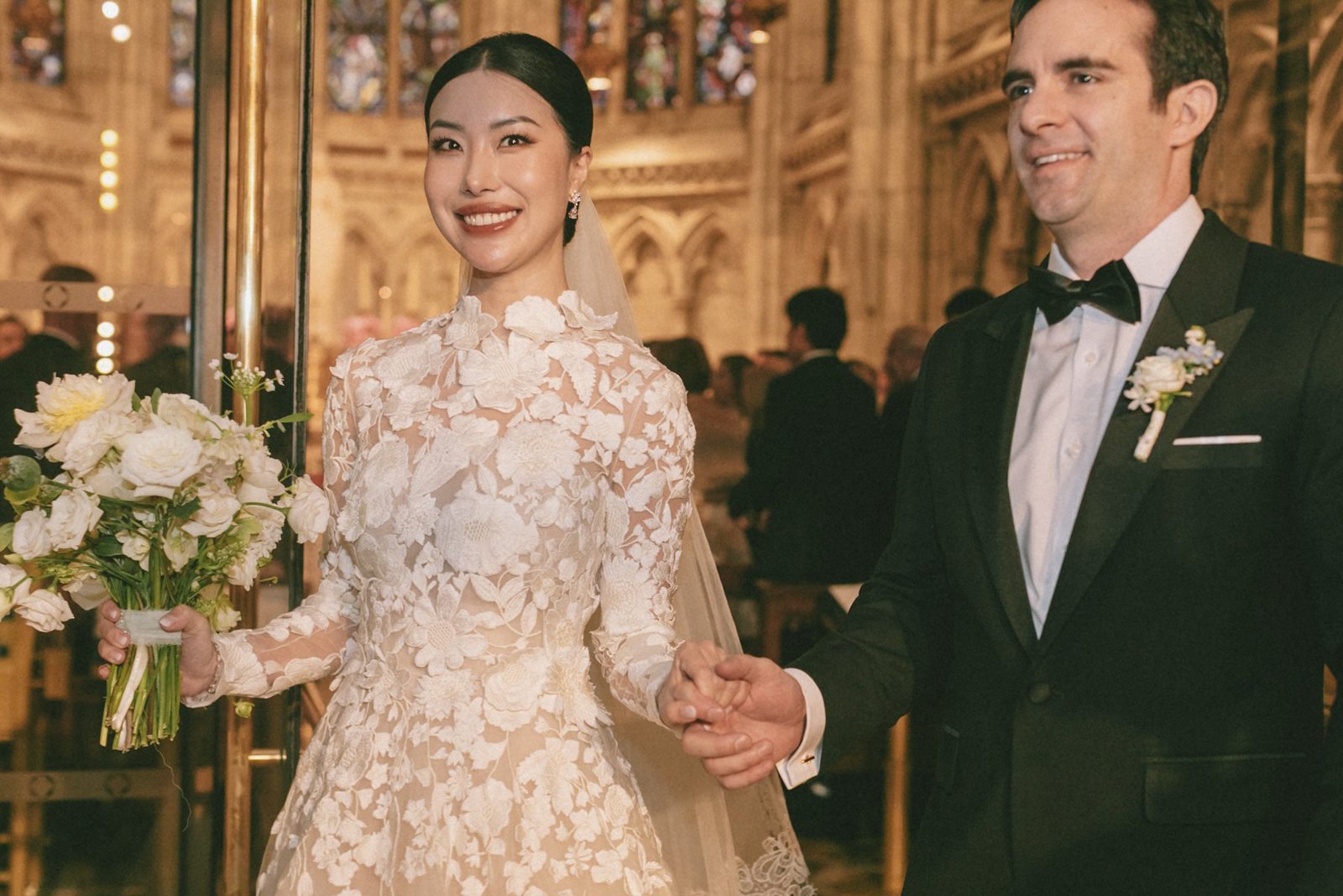 A bride and groom hold hands during their wedding ceremony in an ornate church, with the bride wearing a lace wedding dress holding a bouquet of white flowers, and the groom in a black tuxedo with a bow tie.
