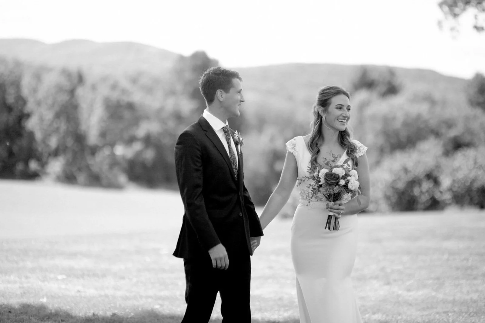 A black-and-white photo of a bride and groom holding hands outdoors during their wedding, with the bride holding a bouquet and both smiling.