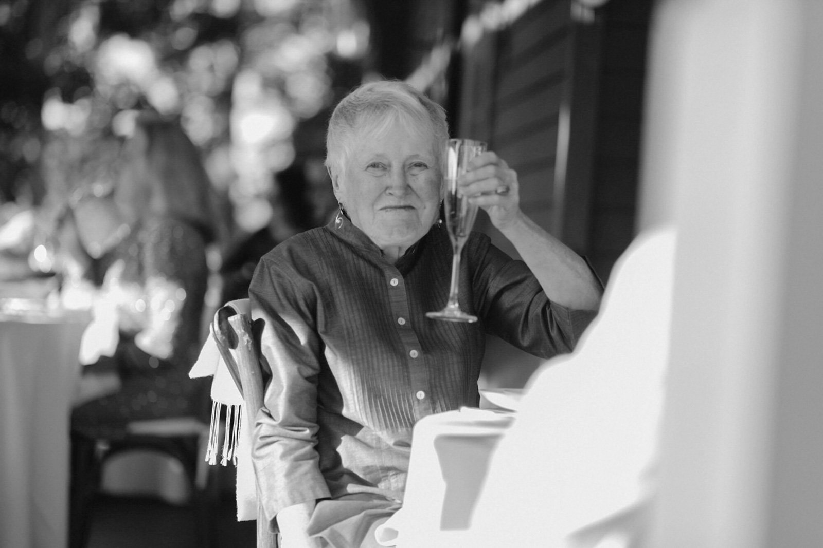An elderly woman sitting at a table, holding a glass of what appears to be champagne, and looking at the camera with a slight smile. 