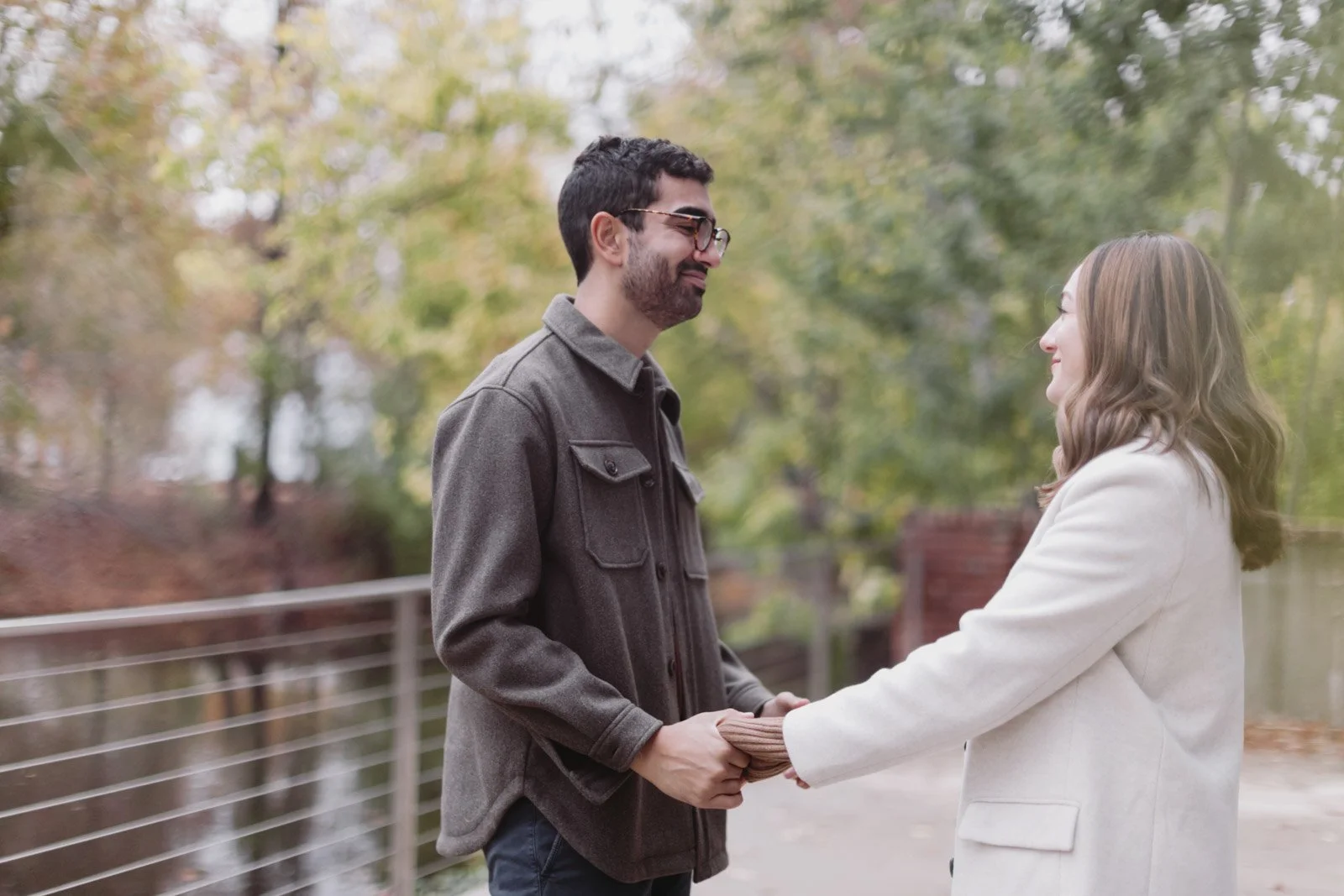 A man and a woman holding hands and smiling at each other outdoors in a park with autumn leaves.
