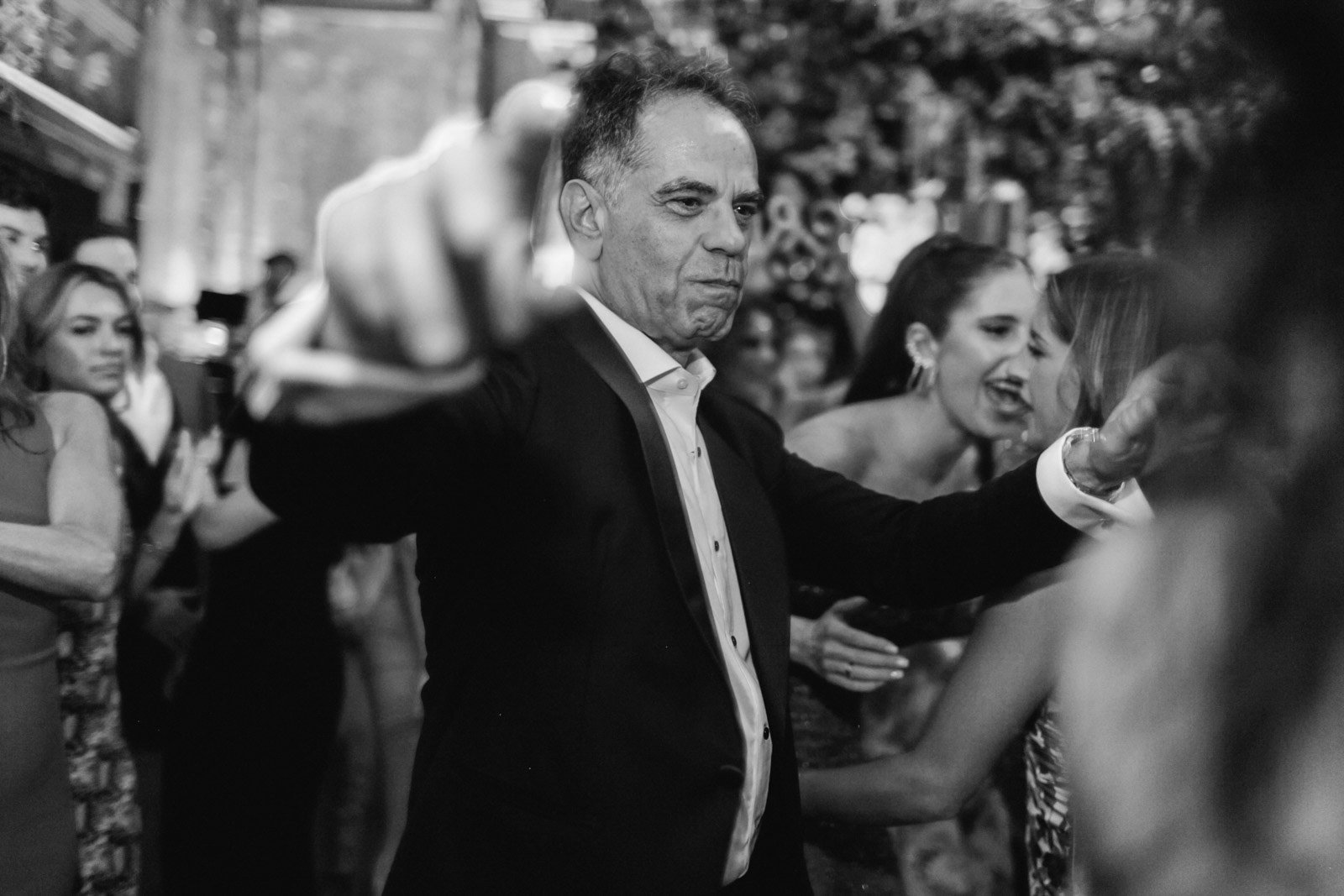 A man in a tuxedo dancing at a crowded social event, with women laughing and dancing in the background.