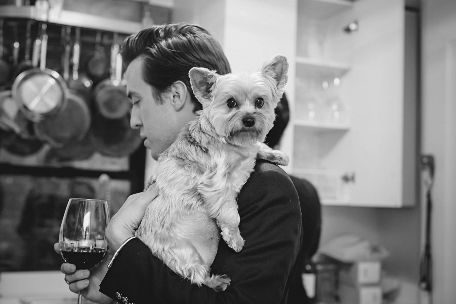 A man with dark hair holding a small dog with light fur, while holding a glass of wine in a kitchen setting in black and white.